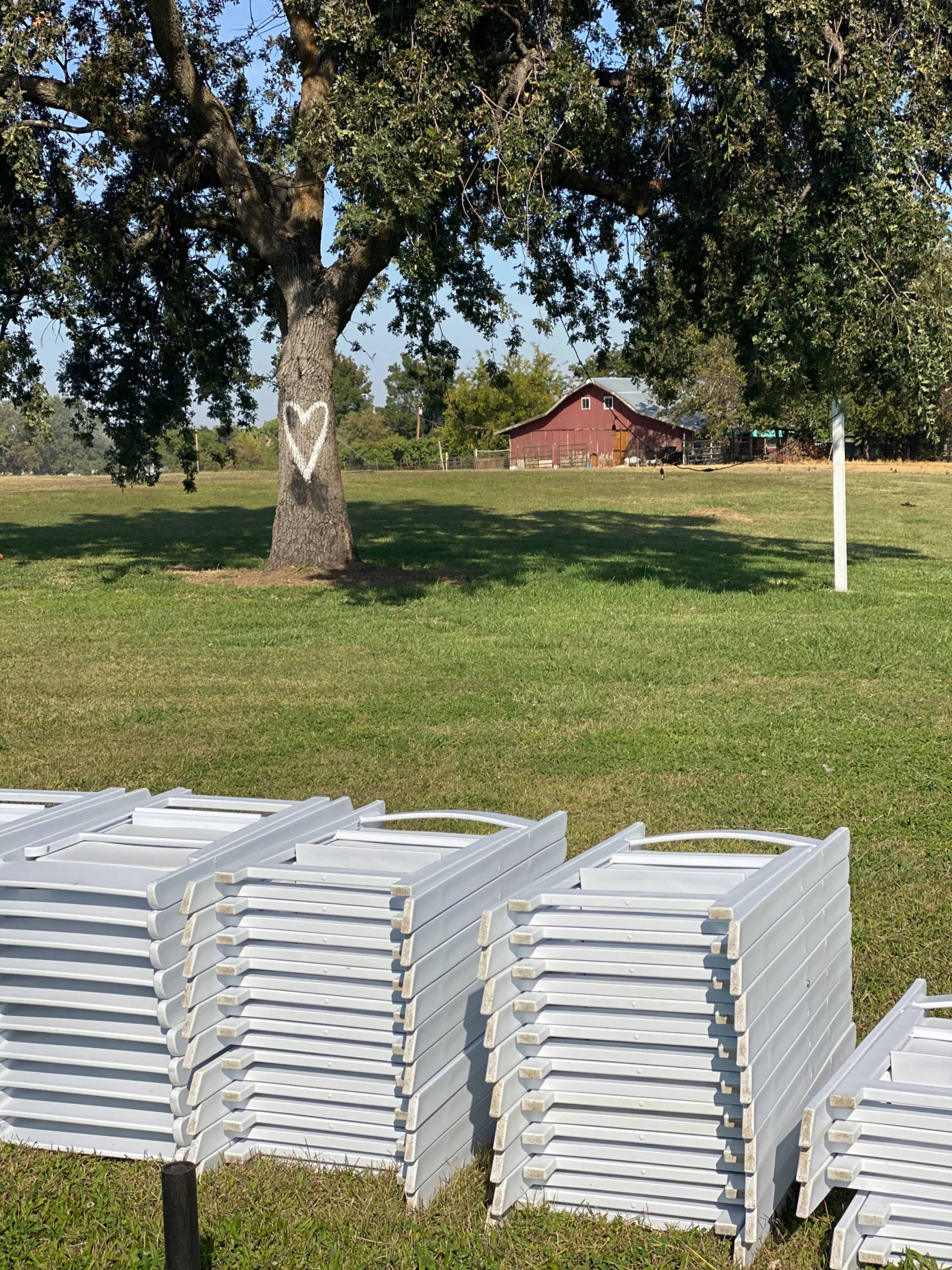 A stack of white, rectangular tables is positioned on the grass in front of a tree with a heart shape carved into its trunk, overlooking a red barn in the background.
