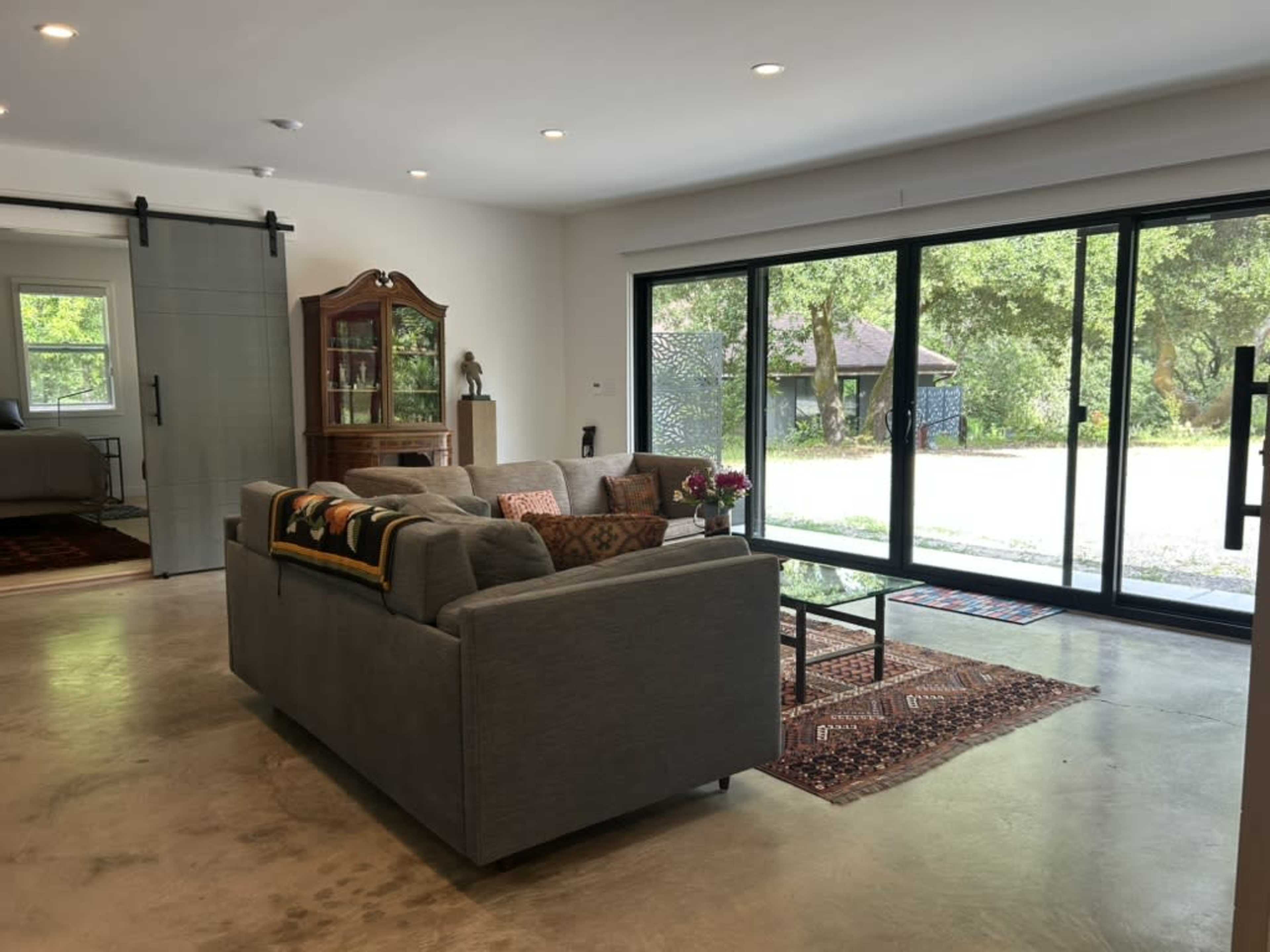 The image shows a modern living room with a gray sofa, a small coffee table, and large glass doors opening to a view of the outdoors.