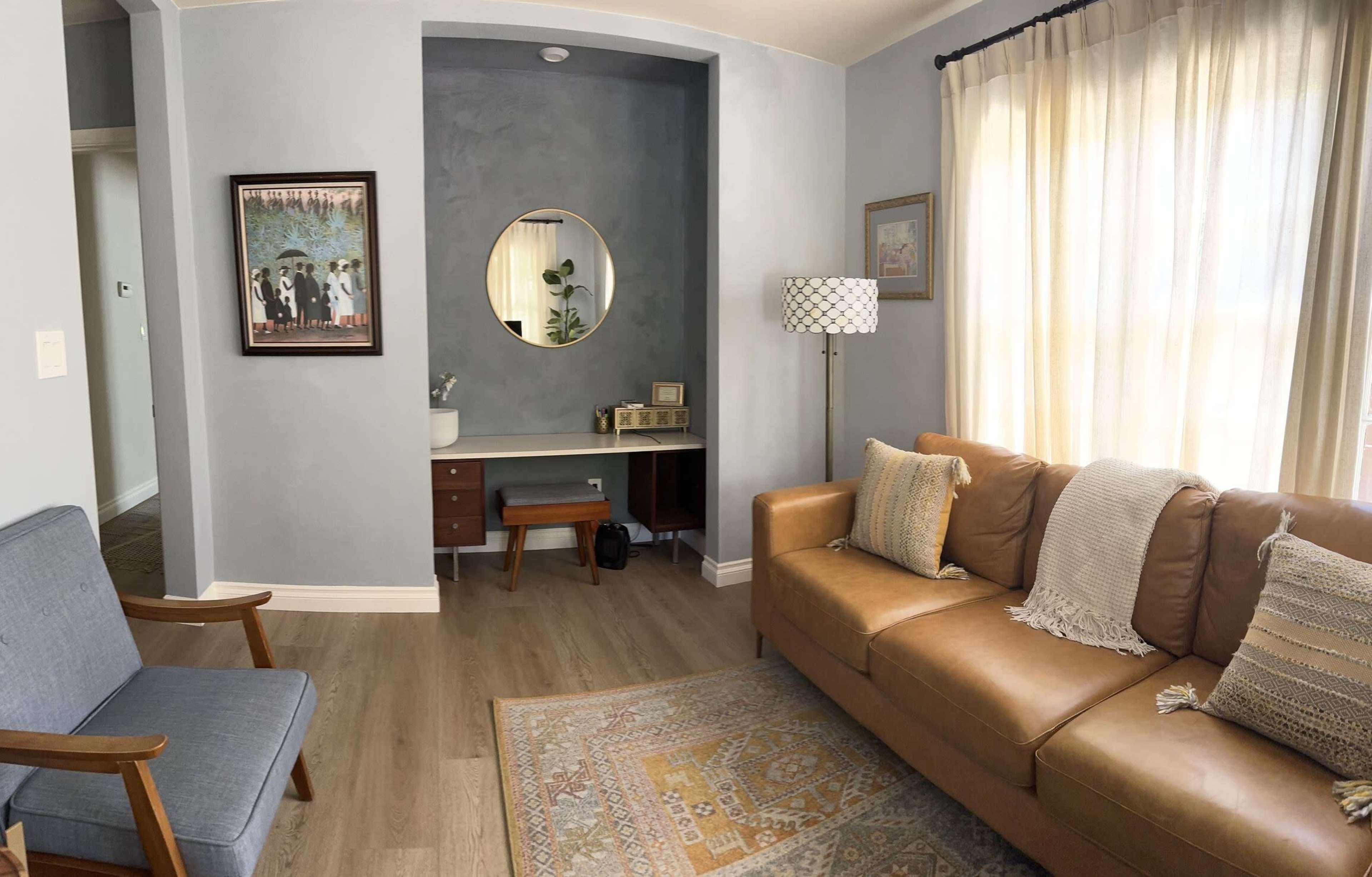 The scene features a cozy living area with a leather sofa, a simple reading chair, and a small workspace illuminated by natural light through sheer curtains.
