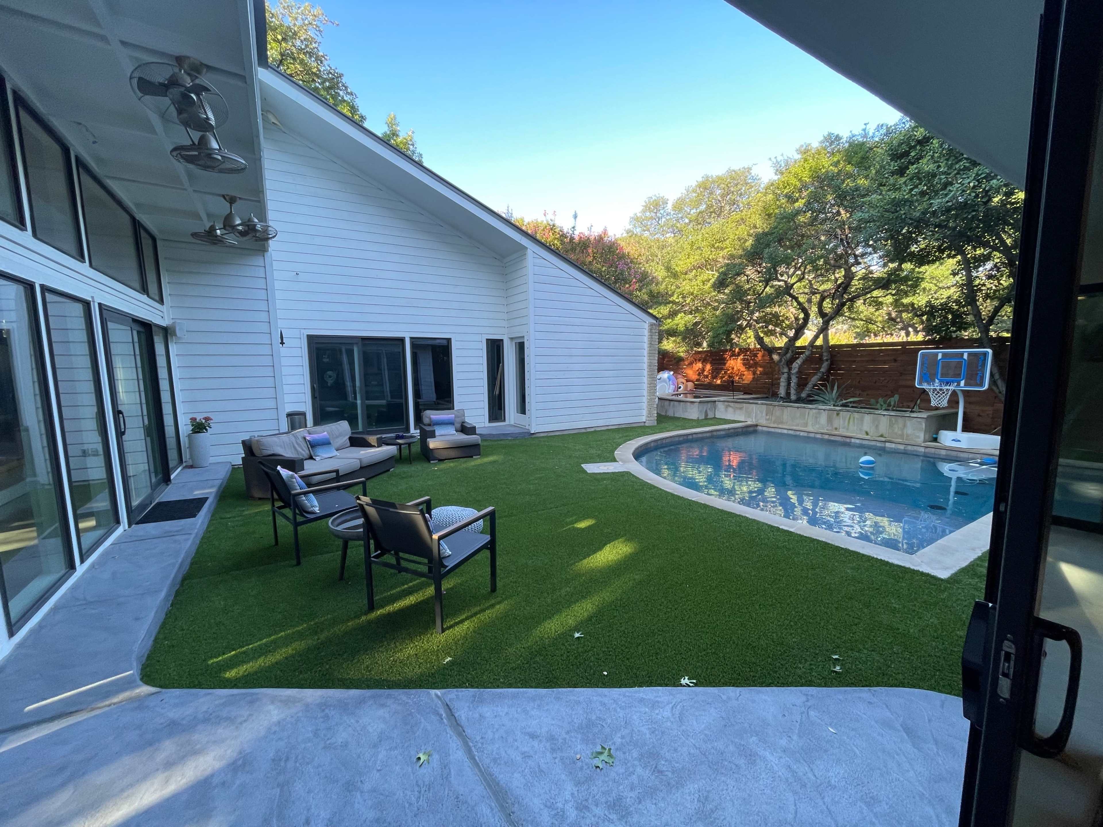 The image shows a modern patio area featuring a swimming pool surrounded by artificial grass and outdoor seating.