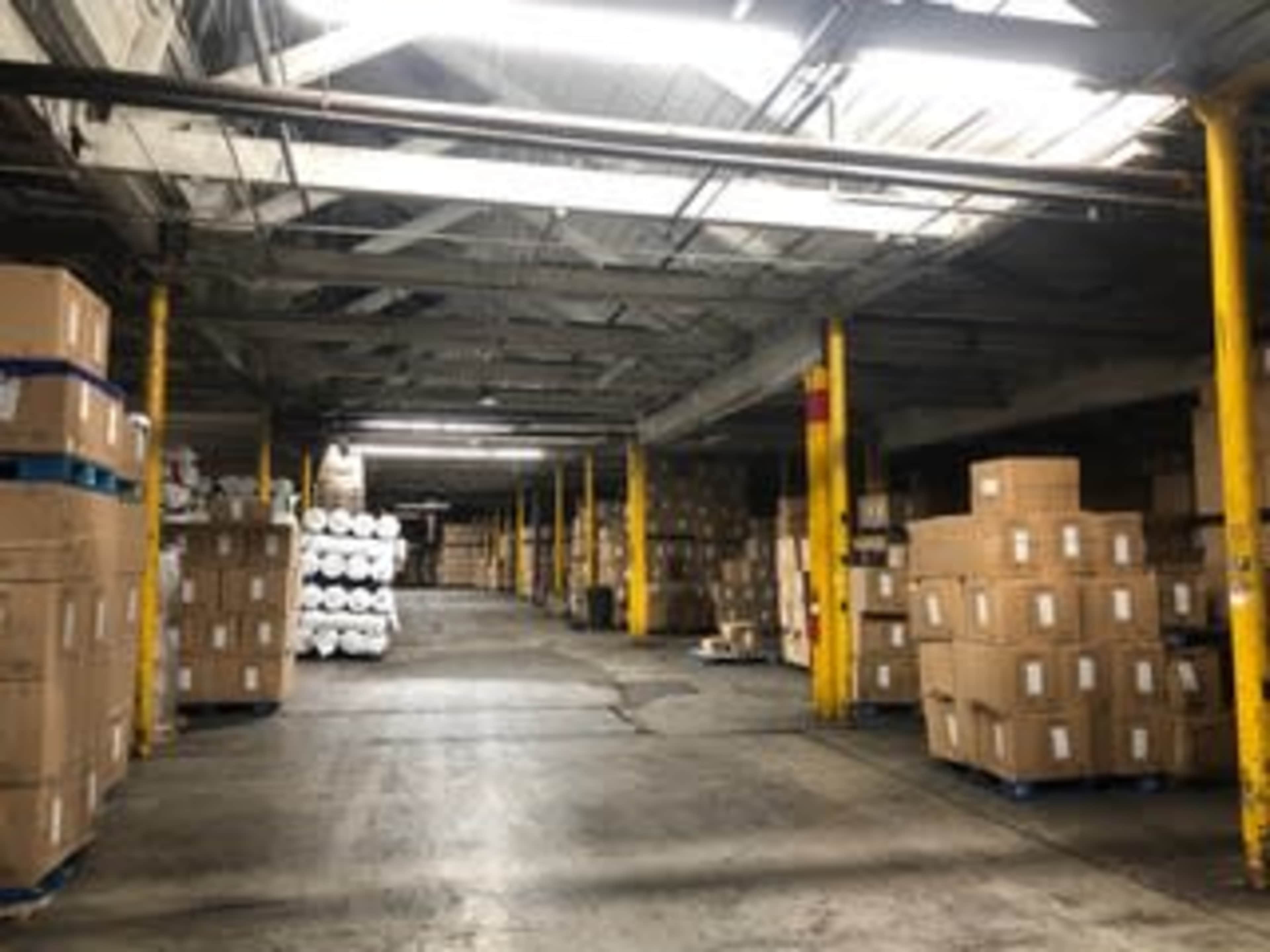 The image shows a large warehouse interior with stacked cardboard boxes and rolls of material lined against the walls.