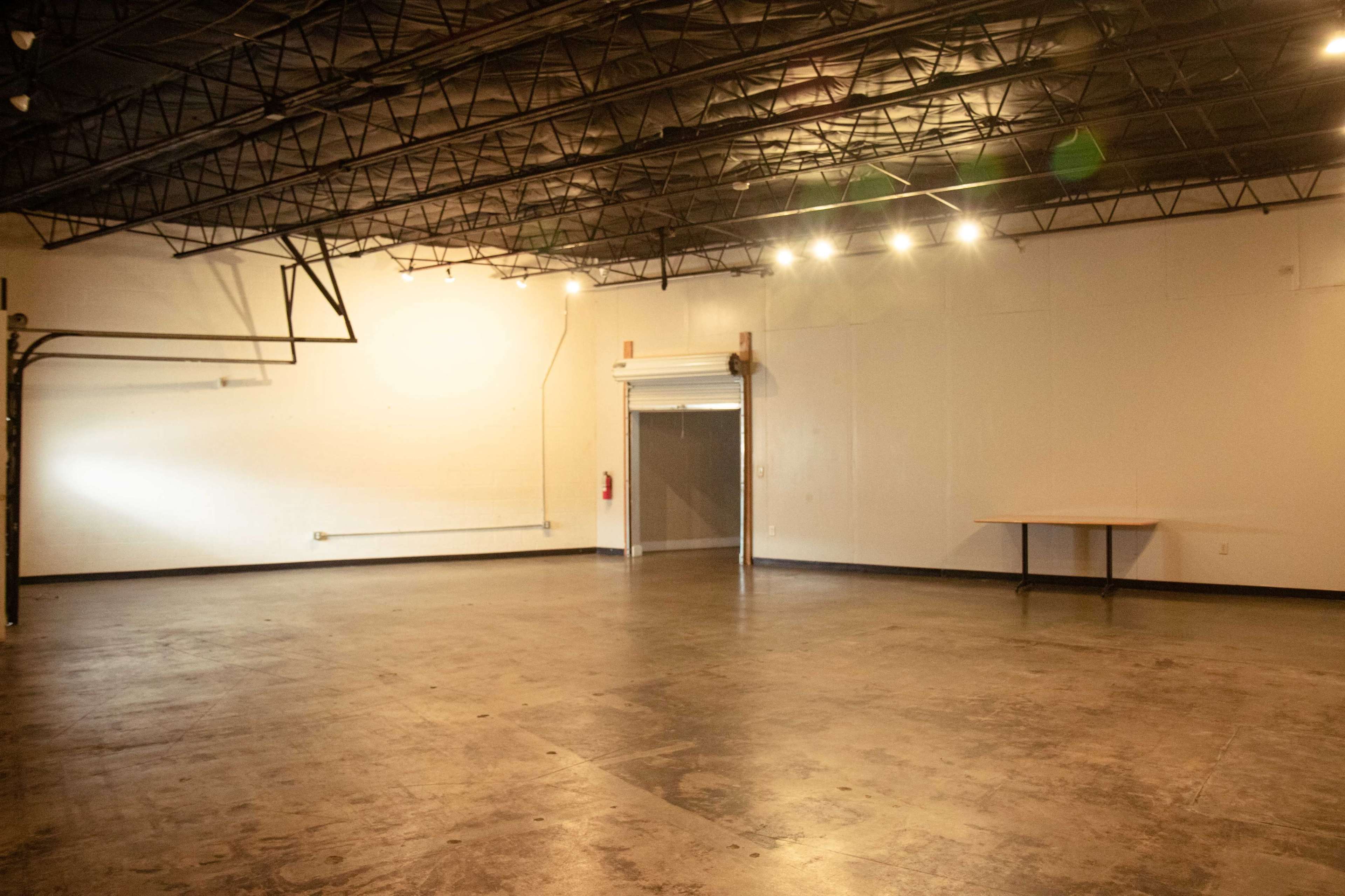 An empty industrial space with a concrete floor, high ceiling, and a table against a wall.