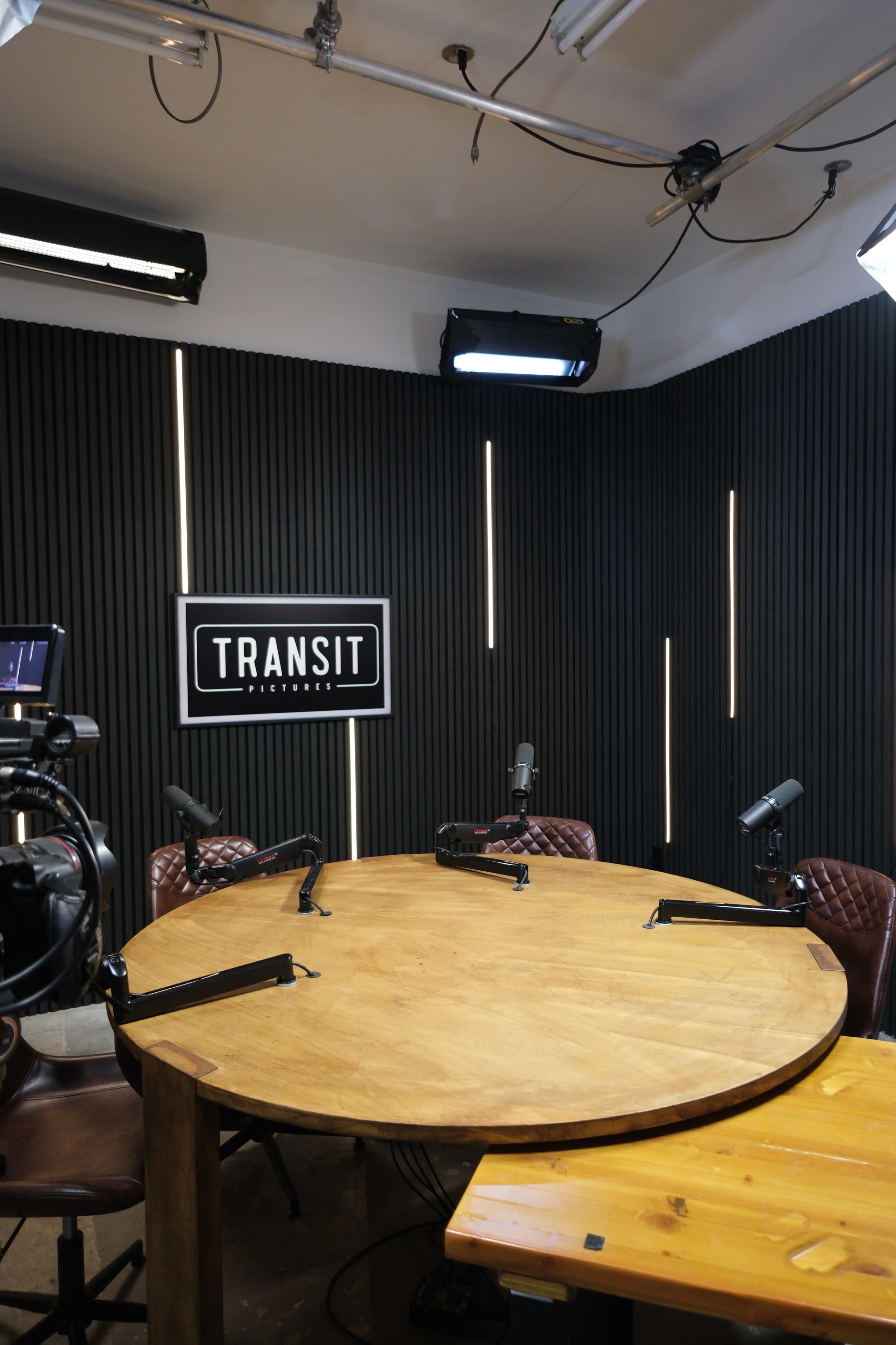 The image shows a podcast studio with a round wooden table, several microphones on stands, and a backdrop with the word "TRANSIT" displayed prominently.