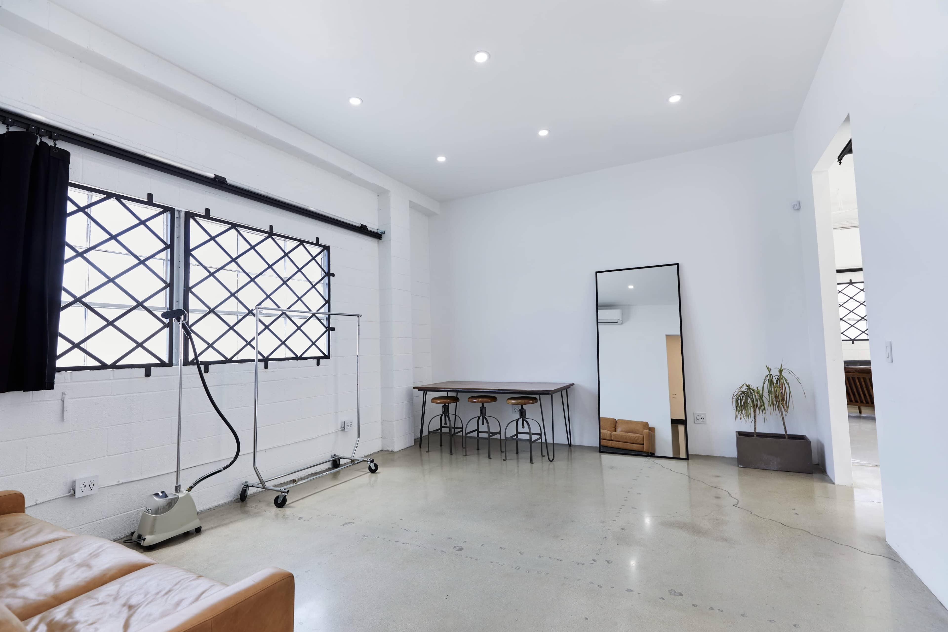 A spacious, minimally furnished room with concrete flooring, a large mirror, a wooden table with stools, and windows featuring decorative metal grilles.
