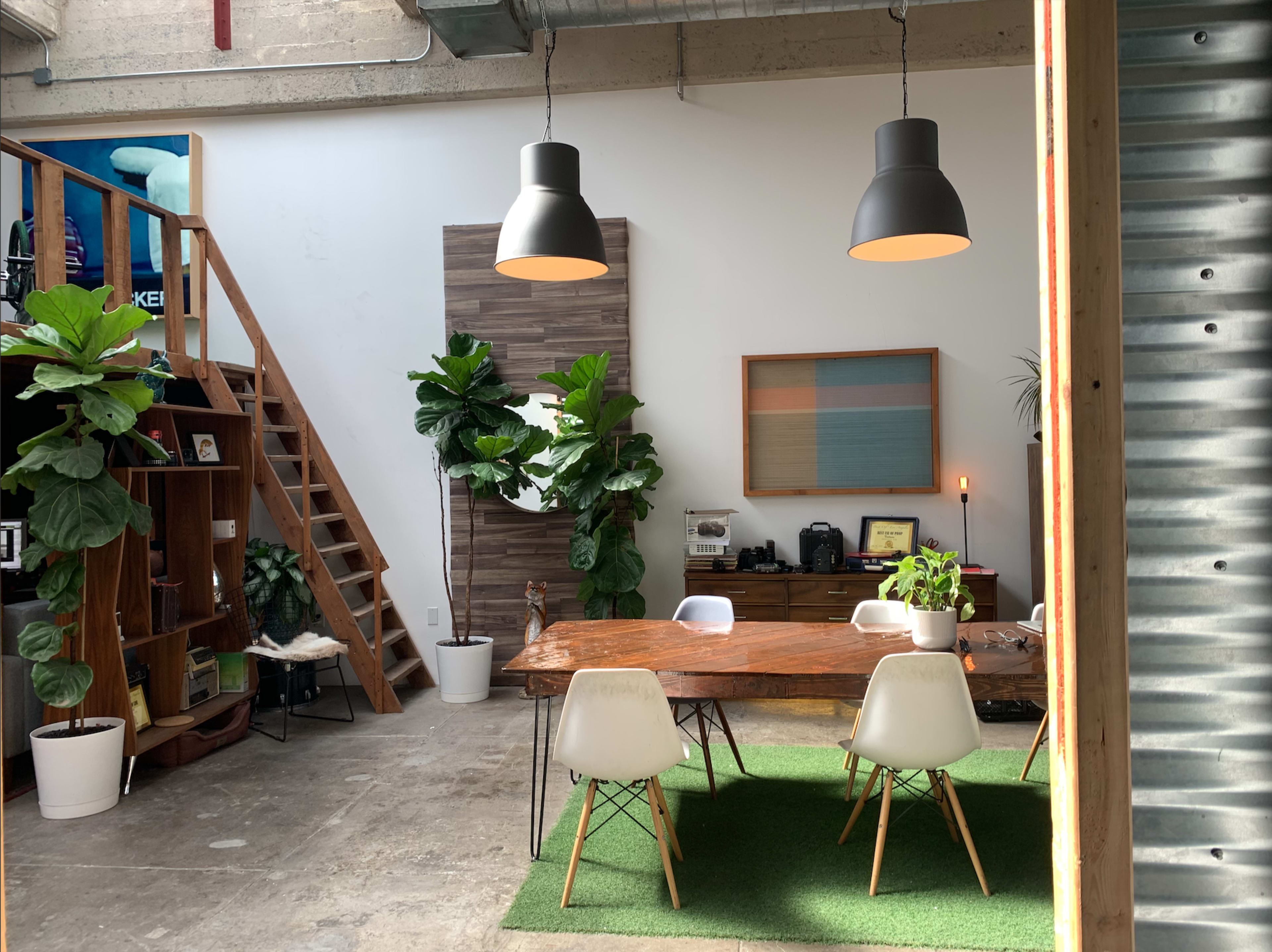 A modern office space features a large wooden table surrounded by white chairs, potted plants, and industrial lighting, with a staircase in the background leading to a loft area.