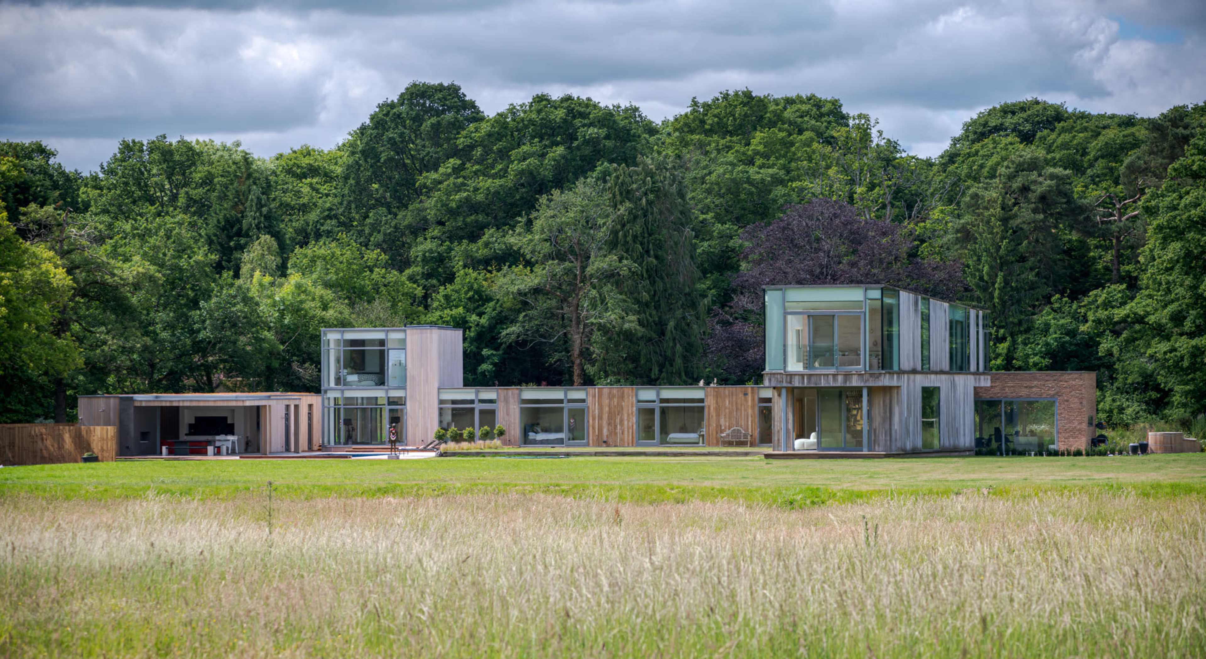 A modern, multi-level house with large glass windows is set against a backdrop of trees and a grassy field.