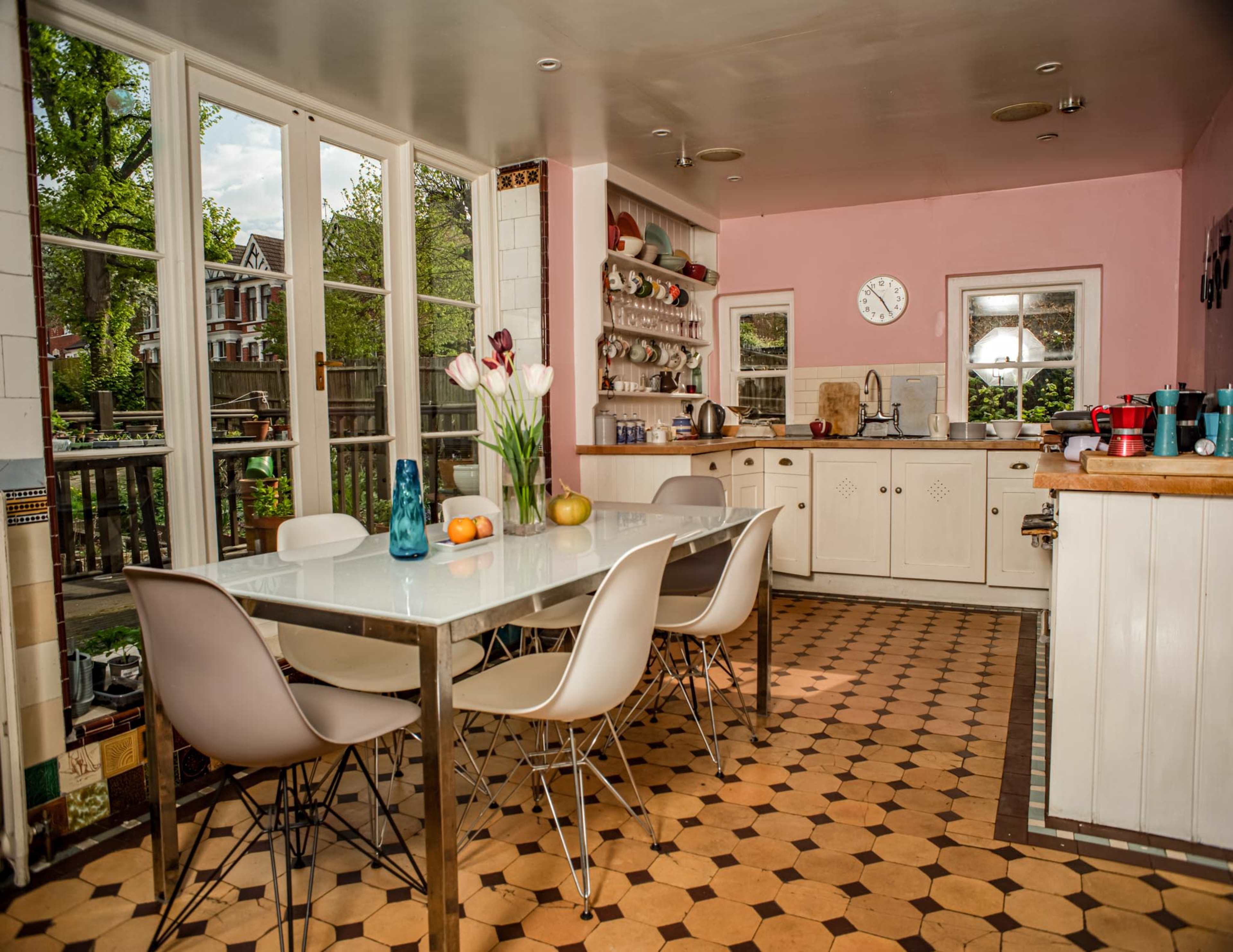 A bright kitchen features a large table surrounded by modern chairs, with dishes and a vase of flowers on the table, and windows letting in natural light.