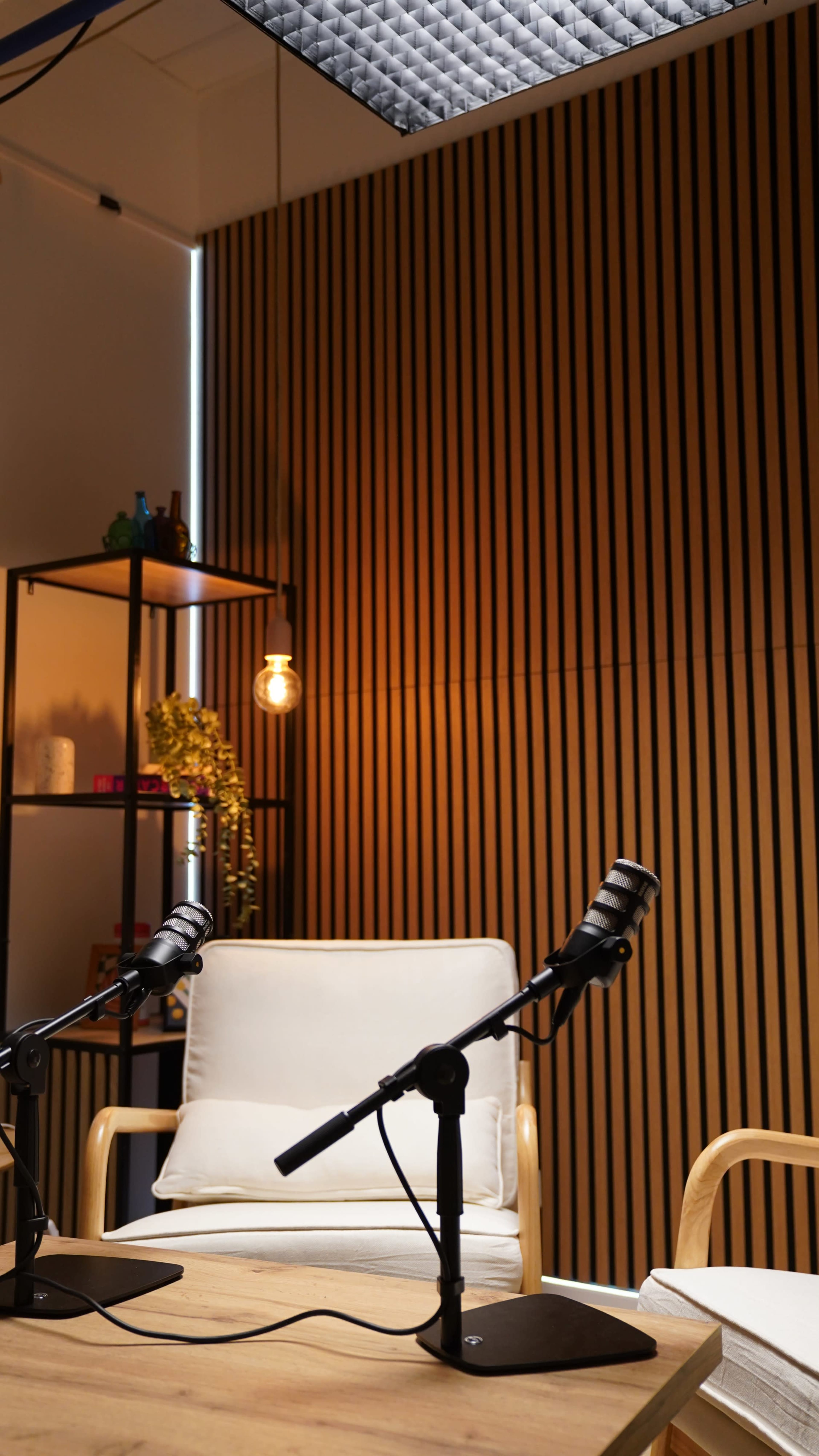 The image shows a modern interview setup with two microphones on stands positioned in front of an empty chair against a backdrop of vertical wooden paneling.