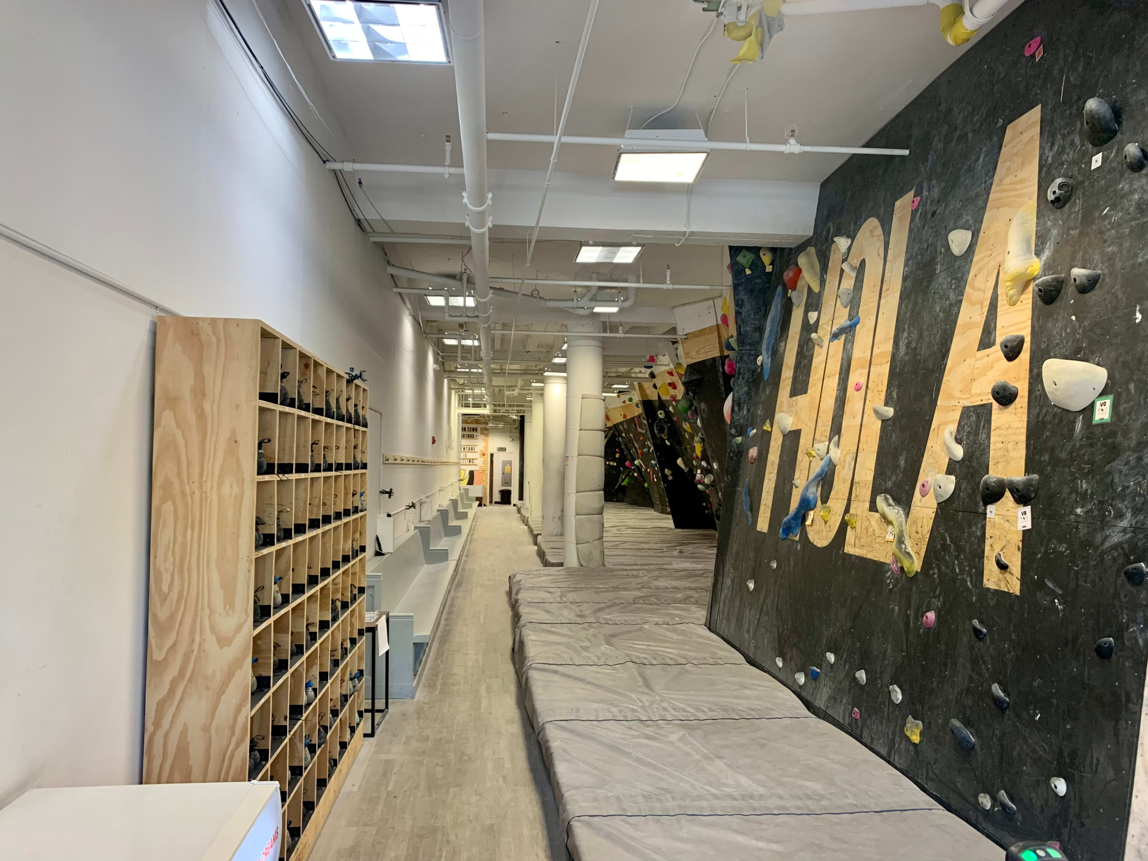 An indoor climbing gym featuring a bouldering wall with colorful holds and padded flooring along a long, brightly lit corridor.