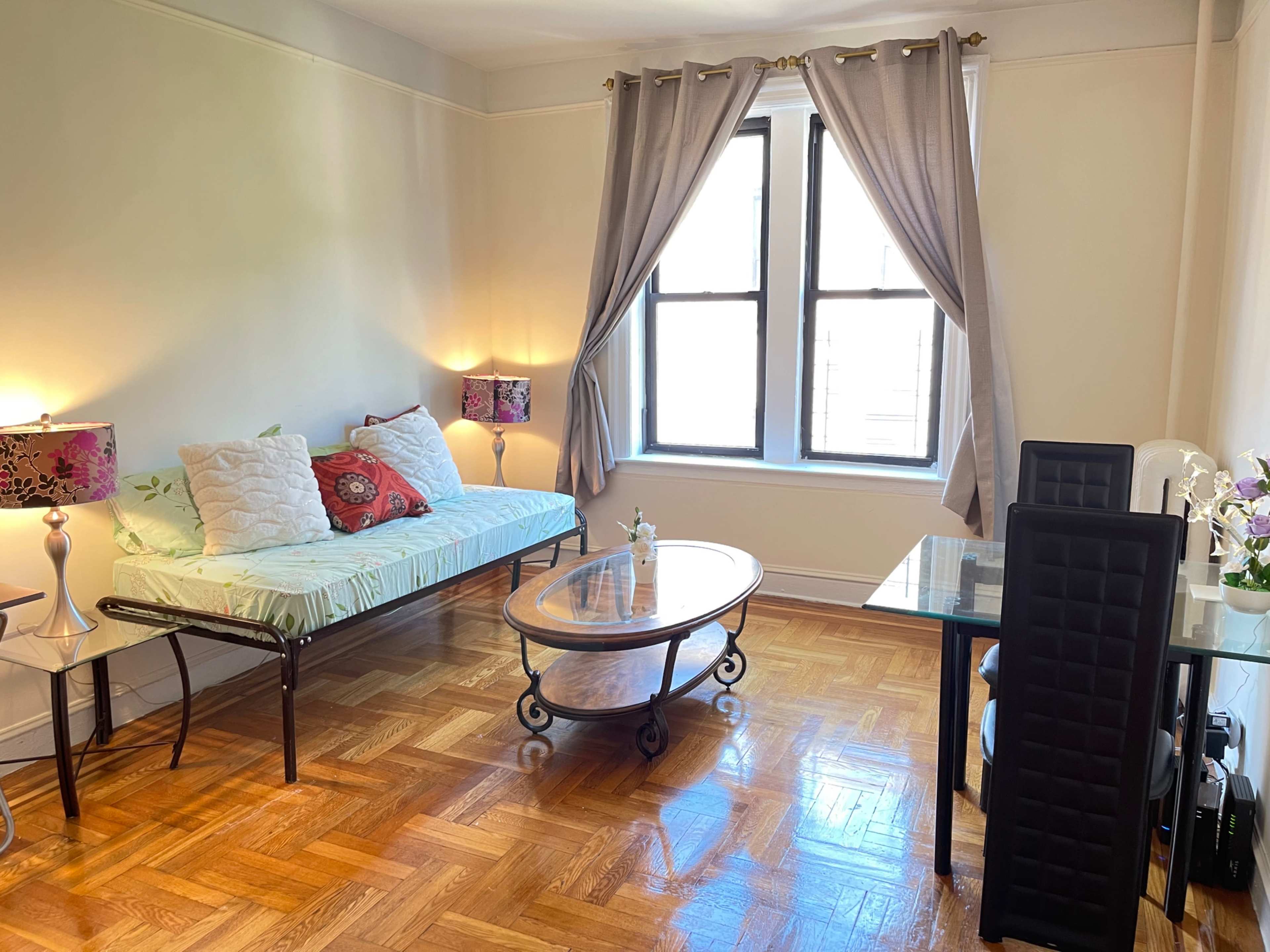 The image shows a small living space with a daybed, a glass coffee table, two chairs, and a window dressed with curtains.