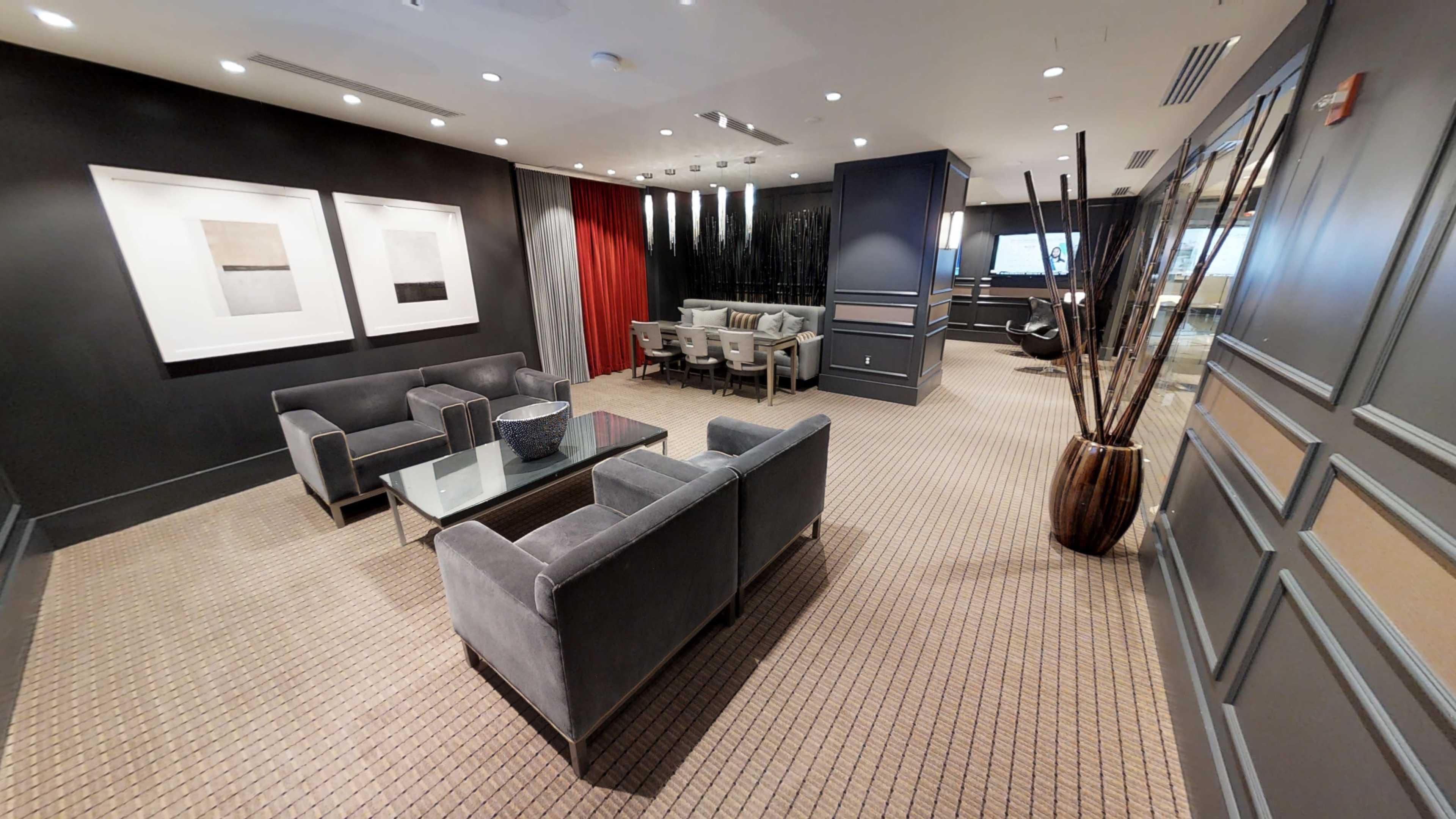 The image shows a modern lounge area with gray sofas, a glass coffee table, a dining table with chairs, and decorative plants against a backdrop of dark walls and a contrasting red curtain.