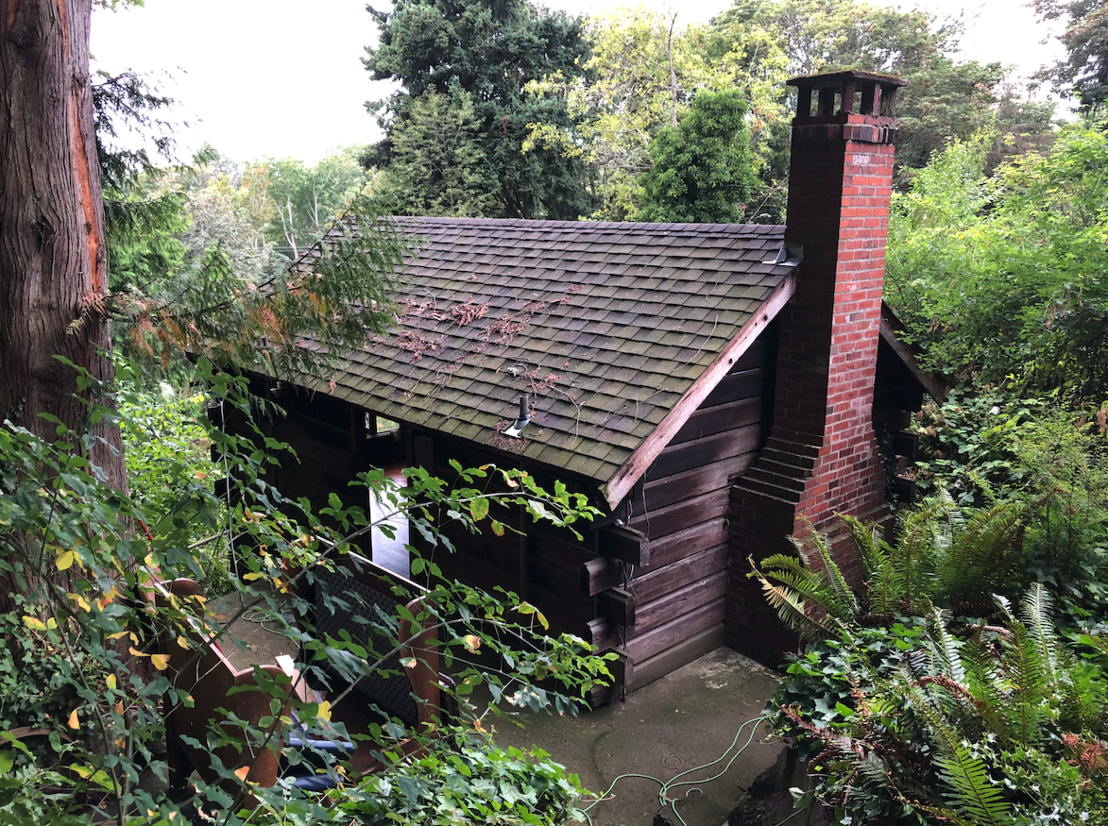 A wooden cabin with a sloped roof and brick chimney is surrounded by lush greenery and trees.