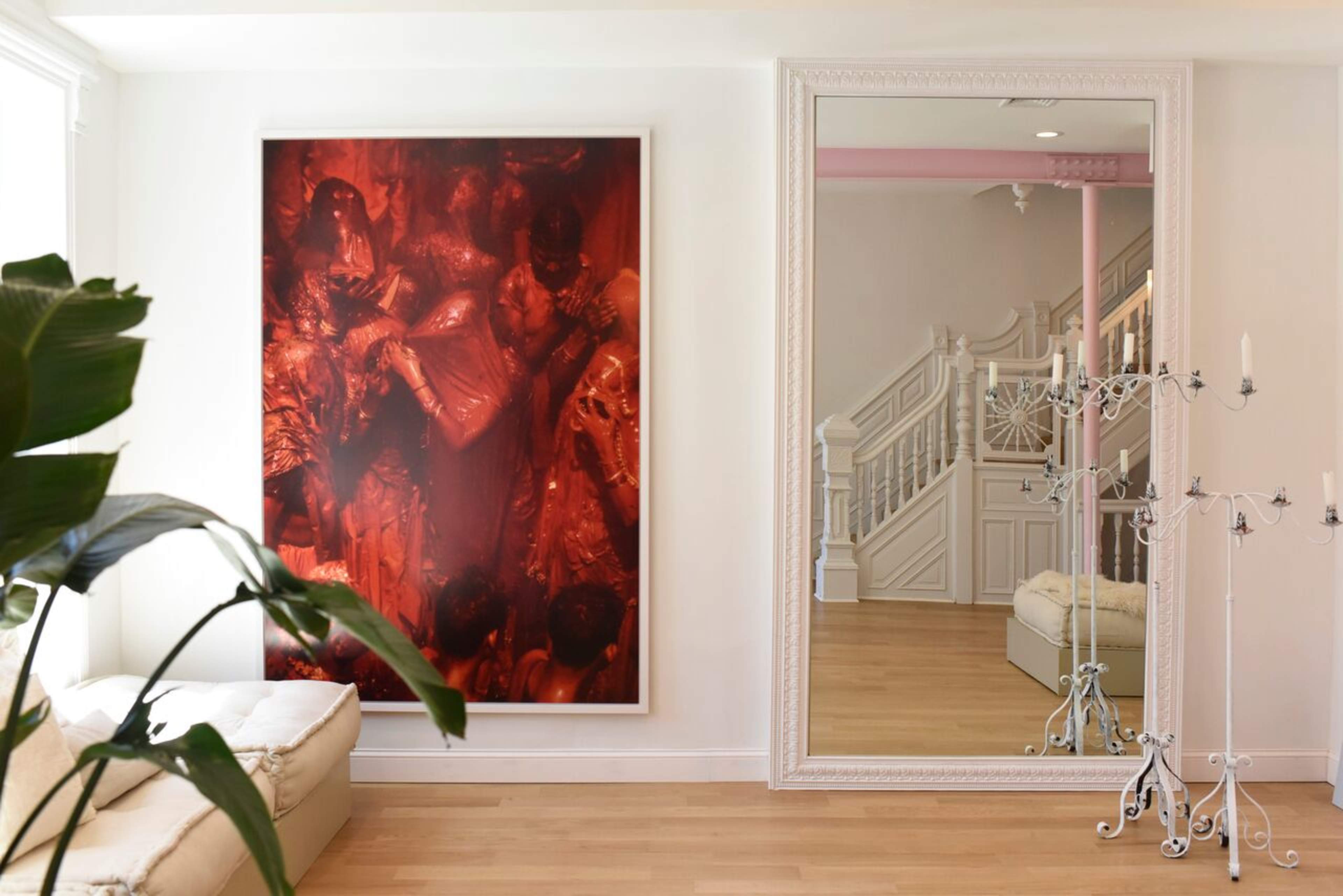 A bright interior with a large red-toned artwork on the wall, a mirror reflecting a staircase, and a decorative candelabra beside a light-colored sofa.