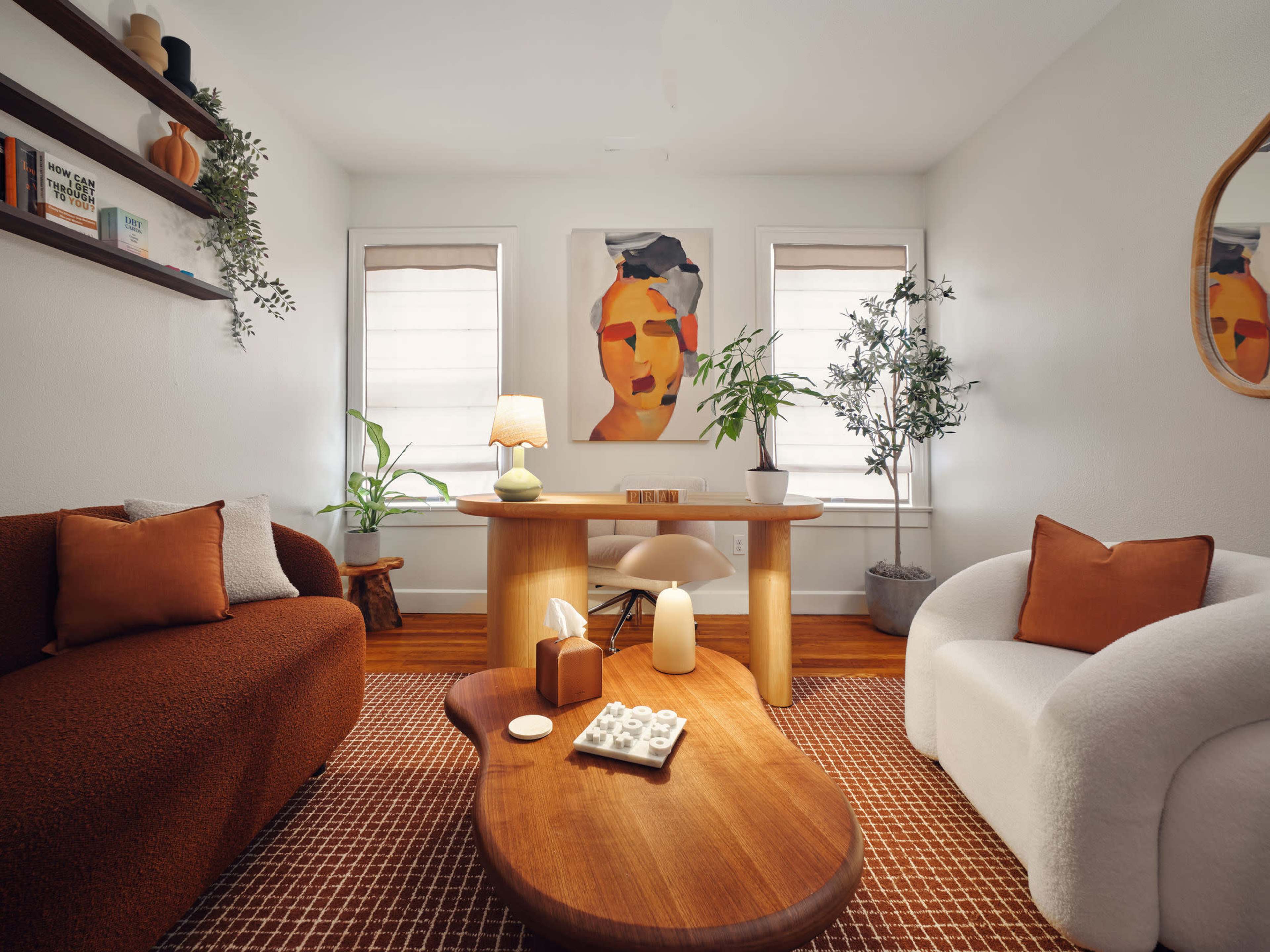 The image shows a cozy living room with two sofas, a wooden coffee table, a desk, and art on the walls, along with plants and decorative items.