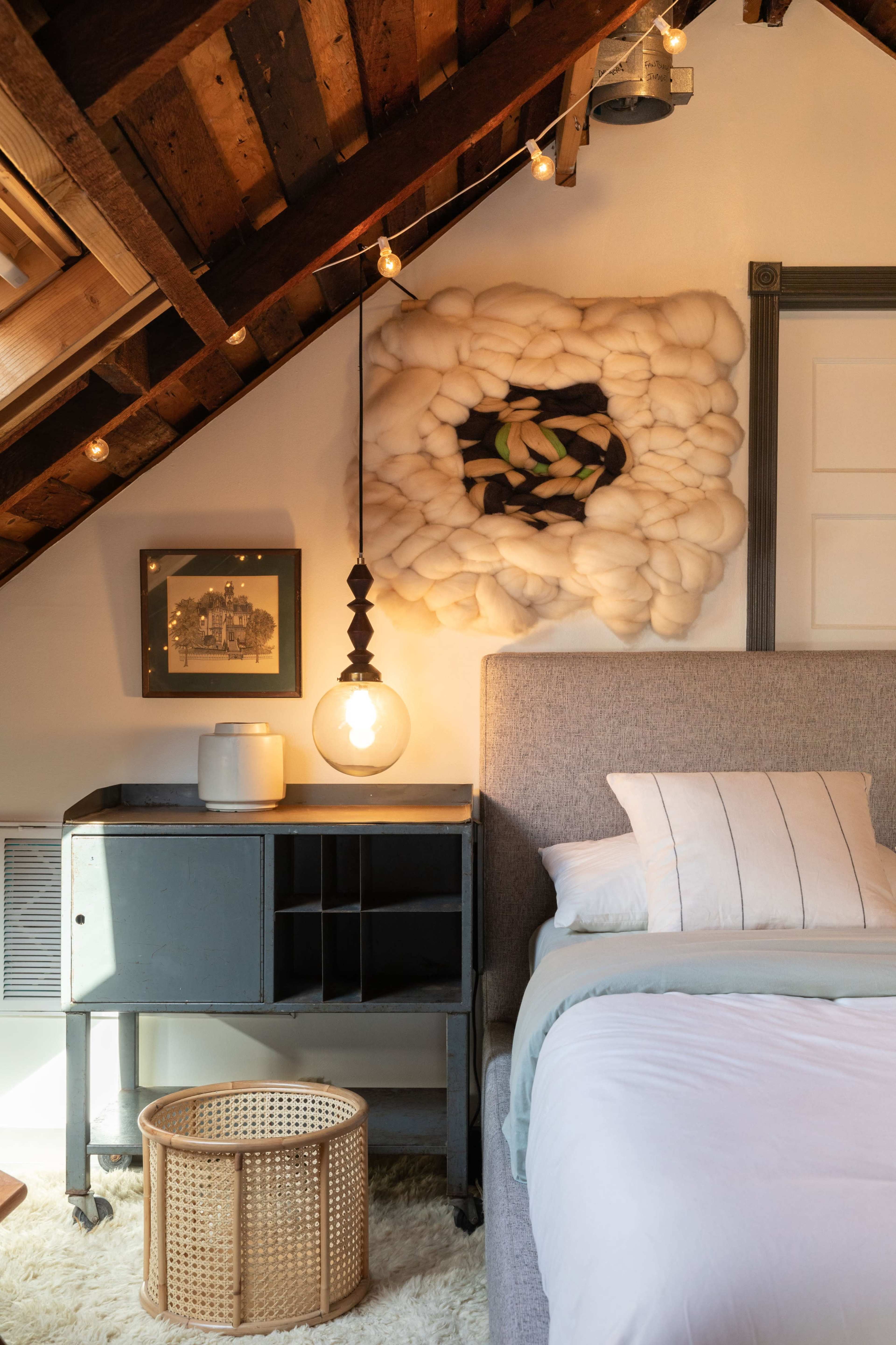 A cozy attic bedroom features a gray upholstered bed, a wooden nightstand with a hanging light, and decorative art on the wall.