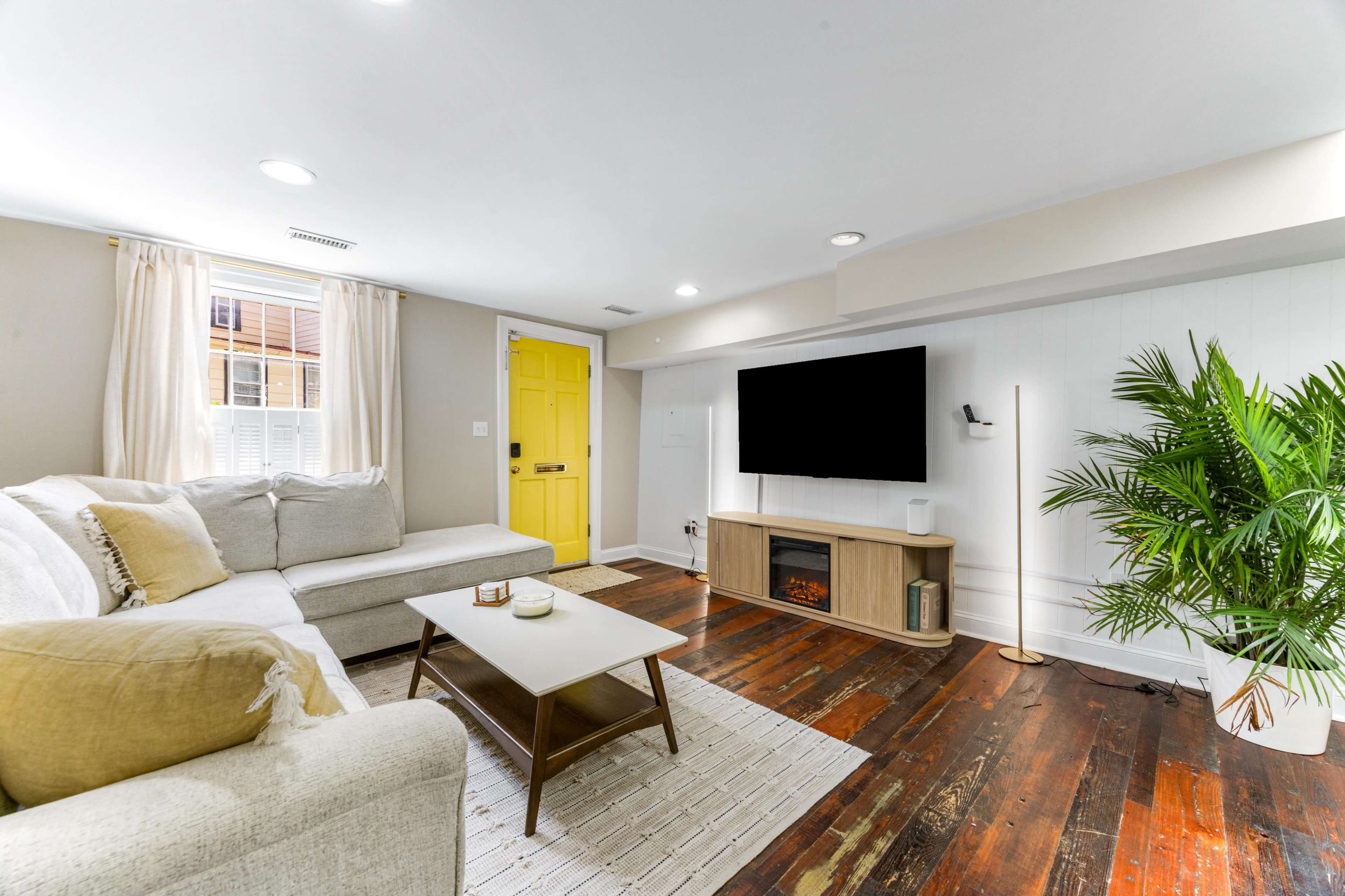 The living room features a light-colored sectional sofa, a wall-mounted television, a decorative plant, and a bright yellow front door.