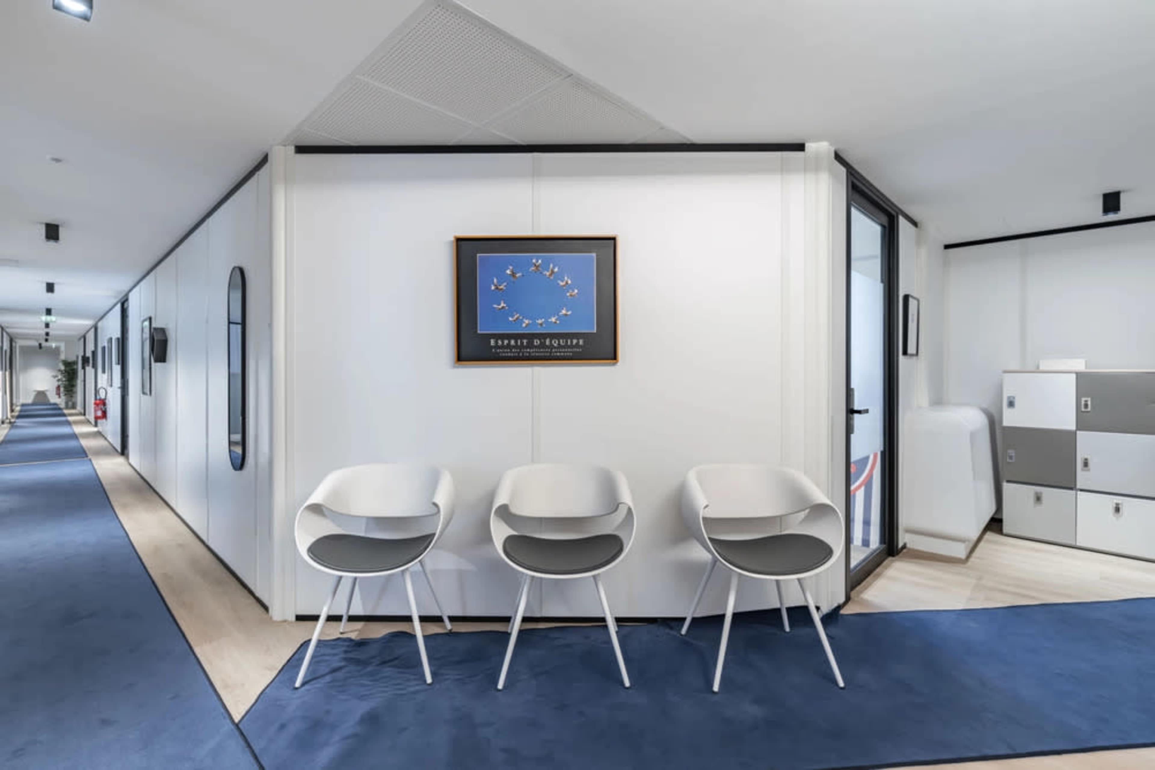 The image shows a modern office interior featuring three white chairs in front of a framed artwork on the wall.