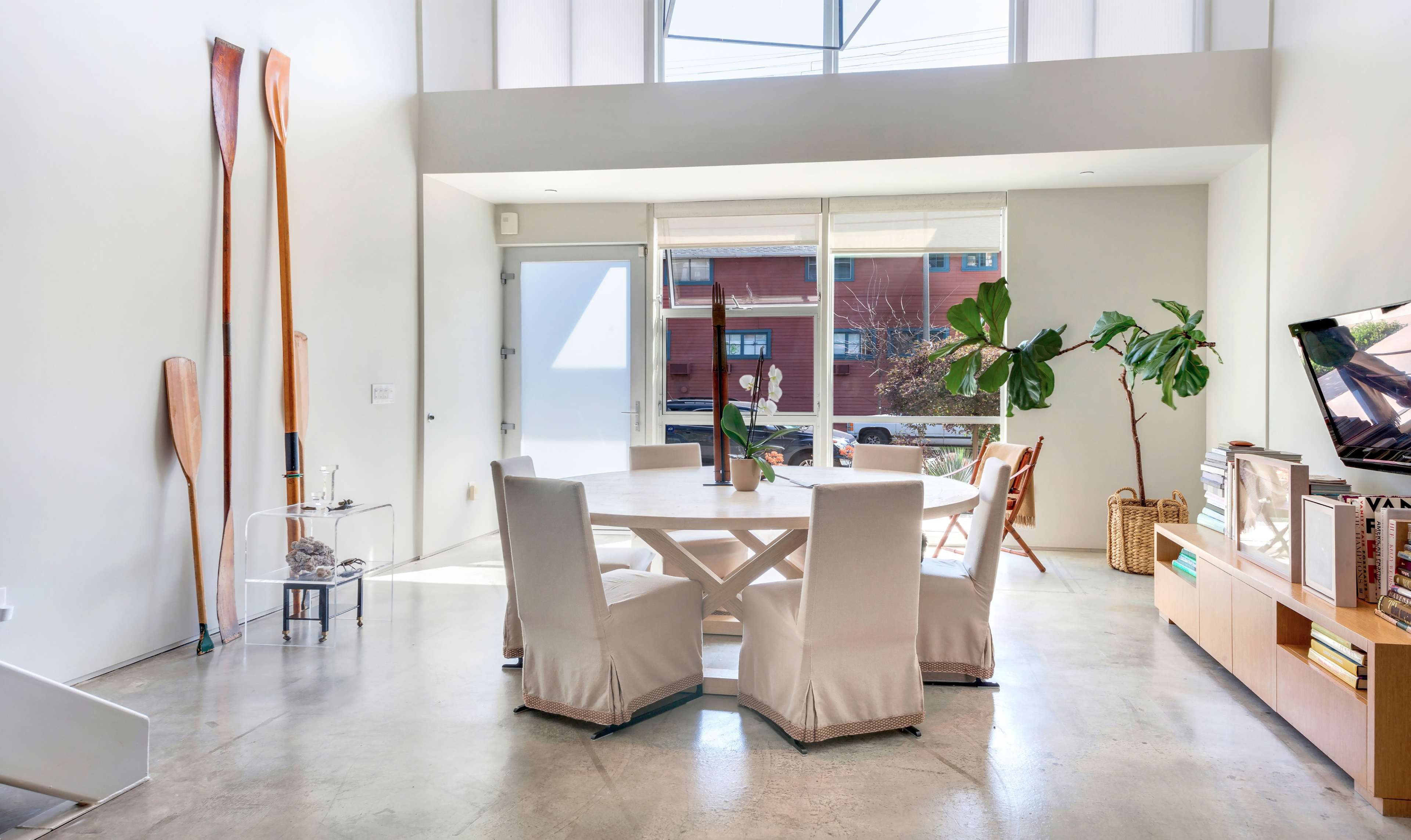 A minimalist dining area features a round table surrounded by four upholstered chairs, with large windows letting in natural light and potted plants nearby.