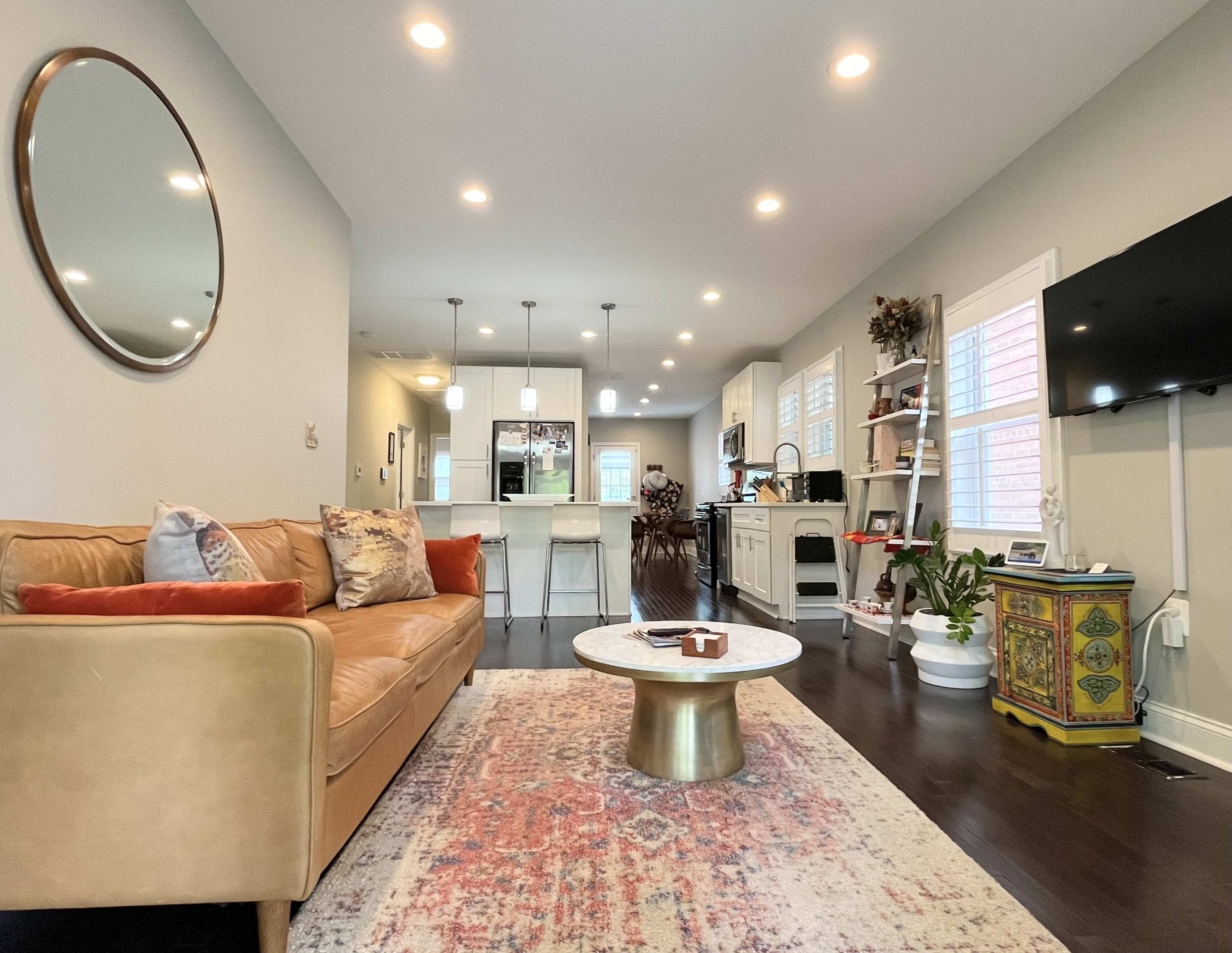A cozy living room features a brown sofa, a round coffee table, and a view of a modern kitchen in the background.
