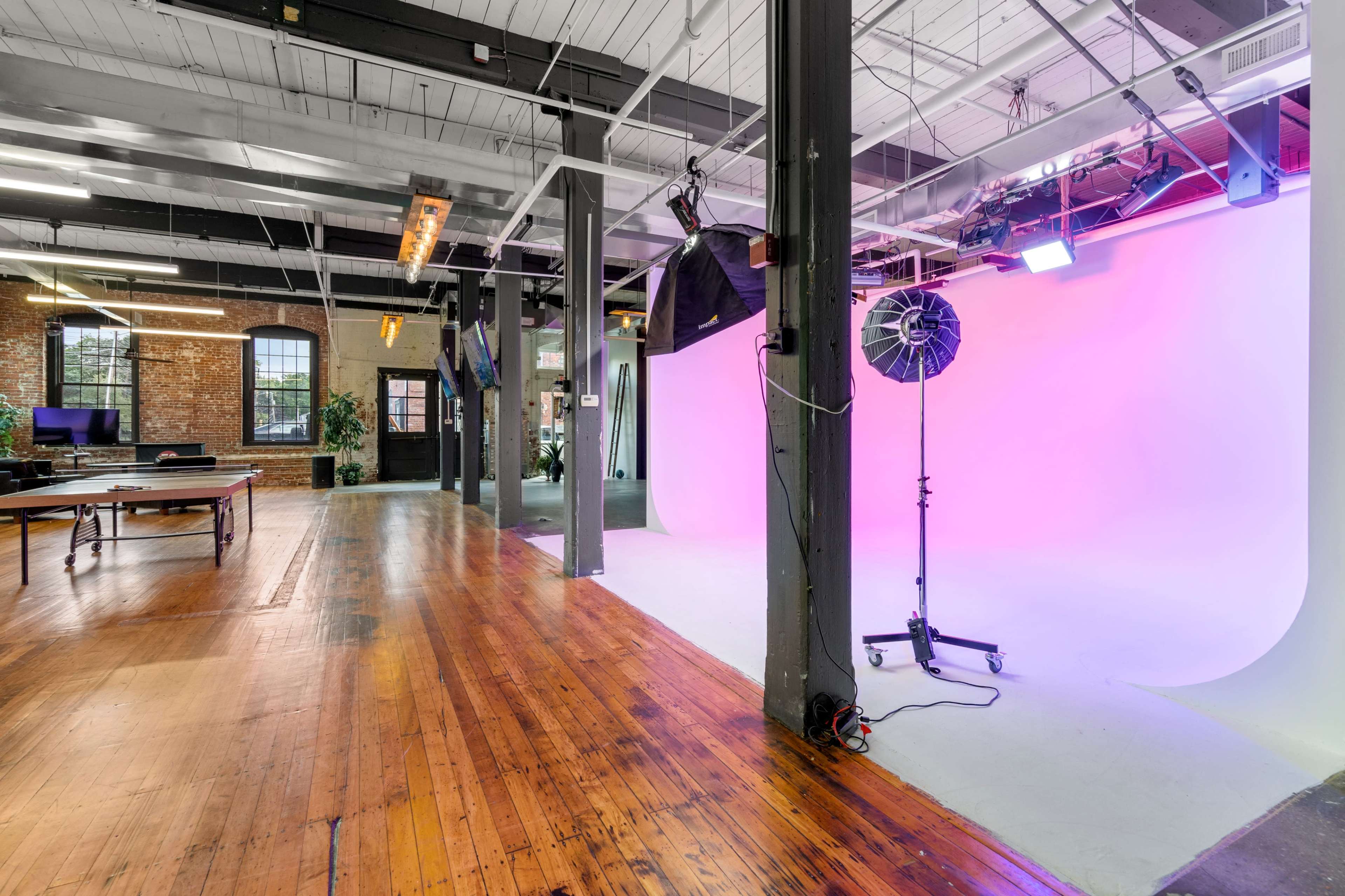 A spacious studio with wooden floors, large windows, and a pink backdrop used for photography, along with studio lights and equipment.