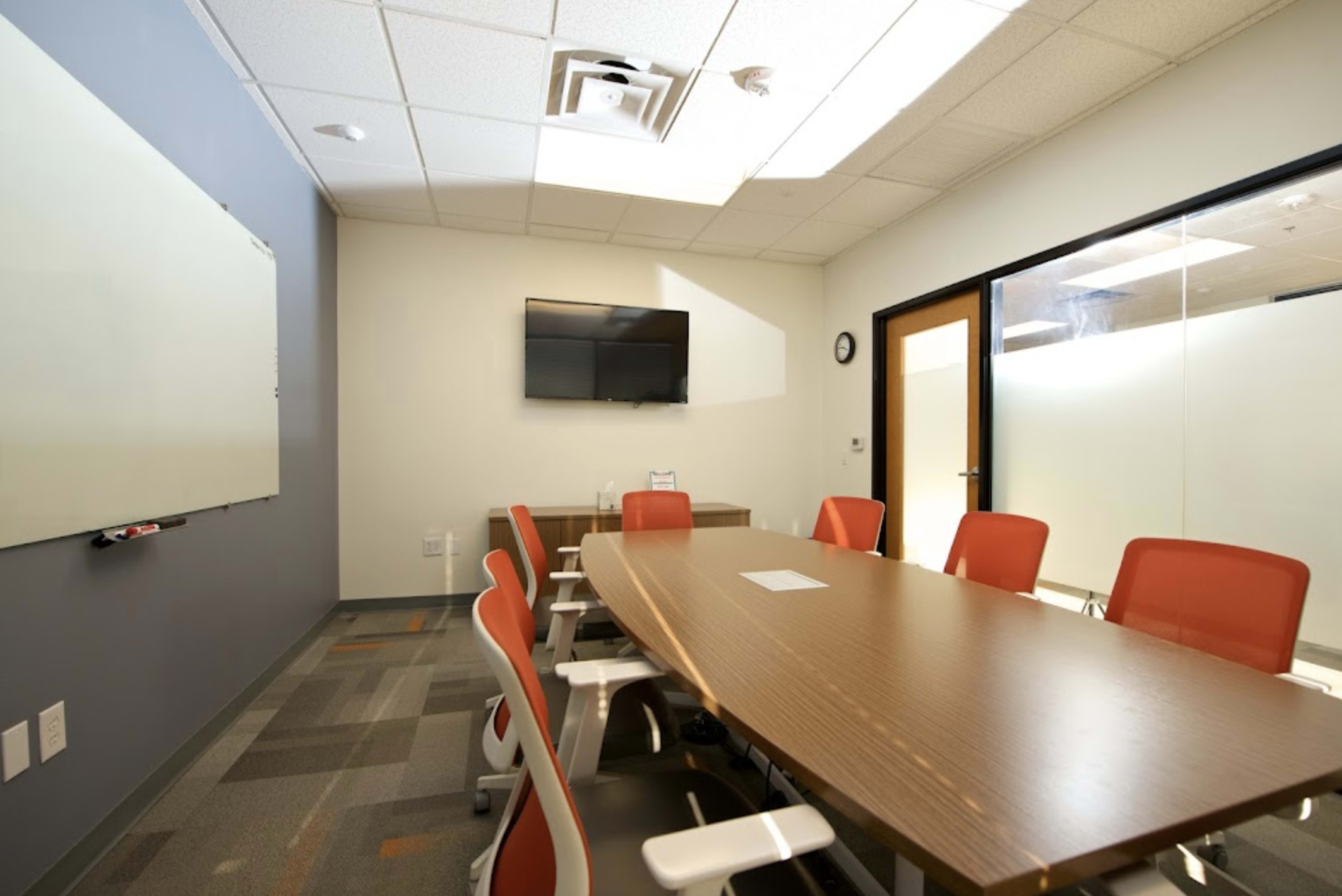 A modern meeting room features a long wooden table surrounded by white chairs, a flat-screen TV mounted on the wall, and a whiteboard.