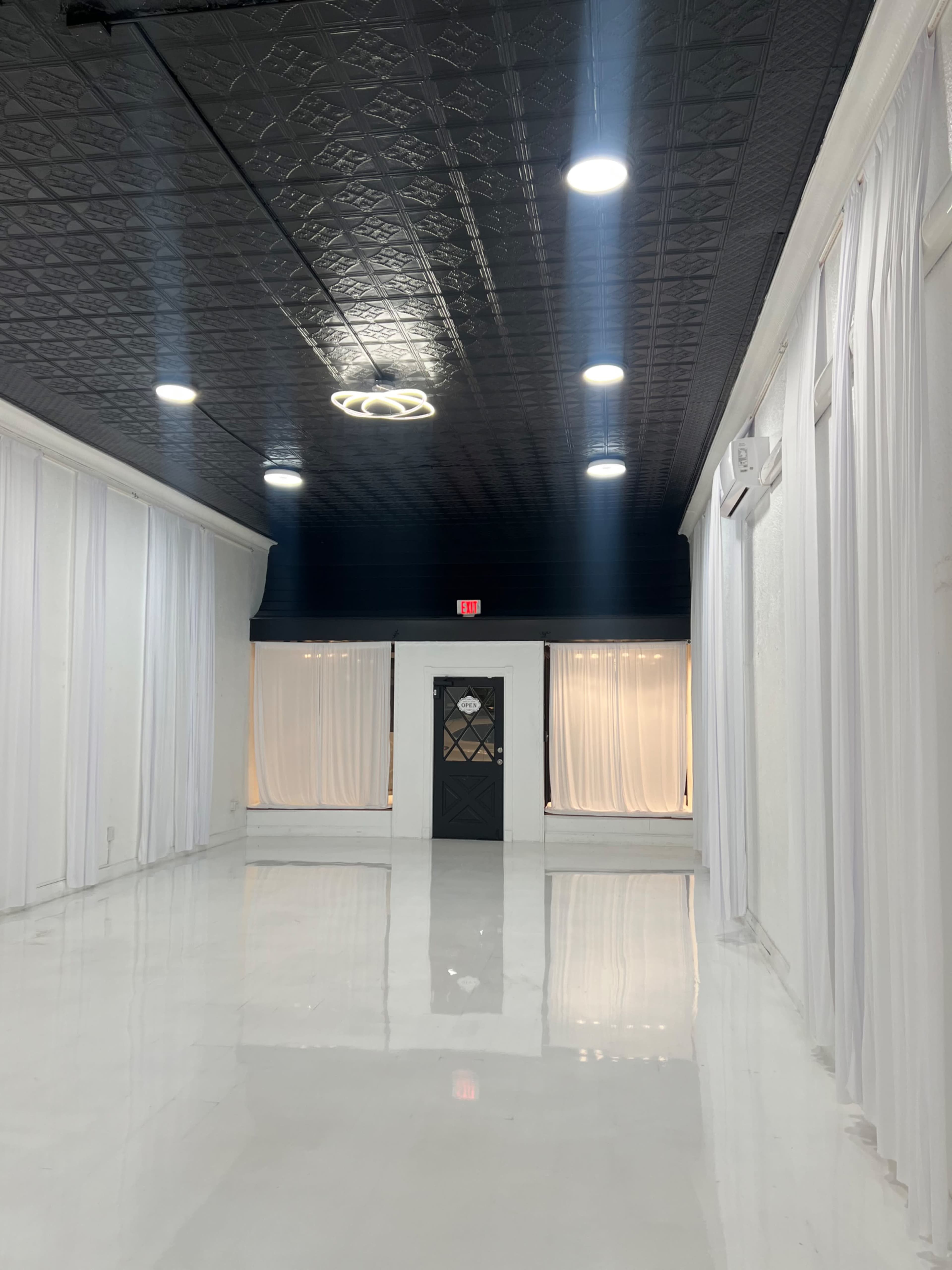 The image shows a spacious, well-lit room with white walls and floor, featuring large, draped curtains and a door at the far end.