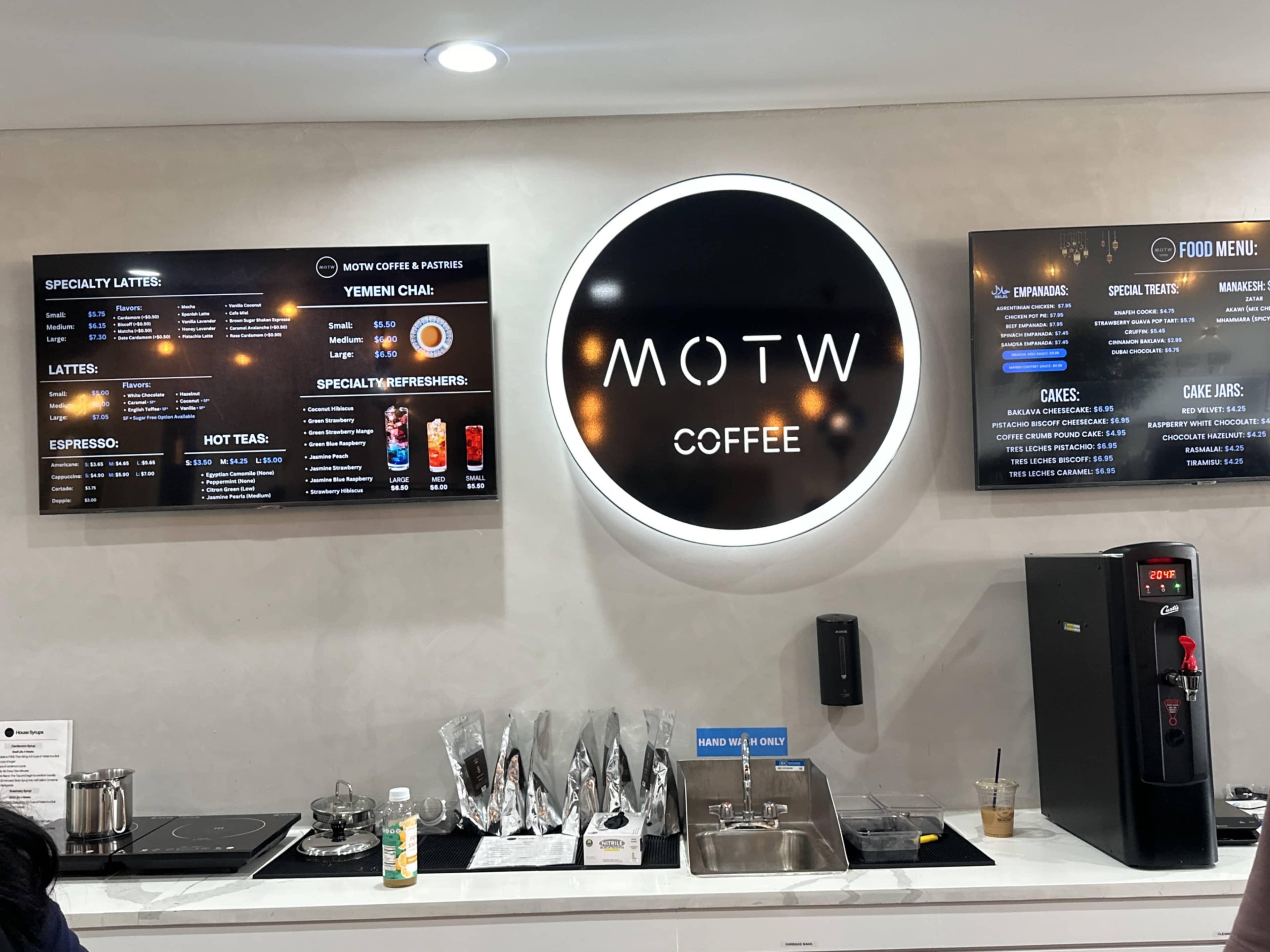 The image shows the interior of a coffee shop featuring a menu board with various beverage options, a circular logo that reads "MOTW COFFEE," and a coffee machine next to a drink preparation area.