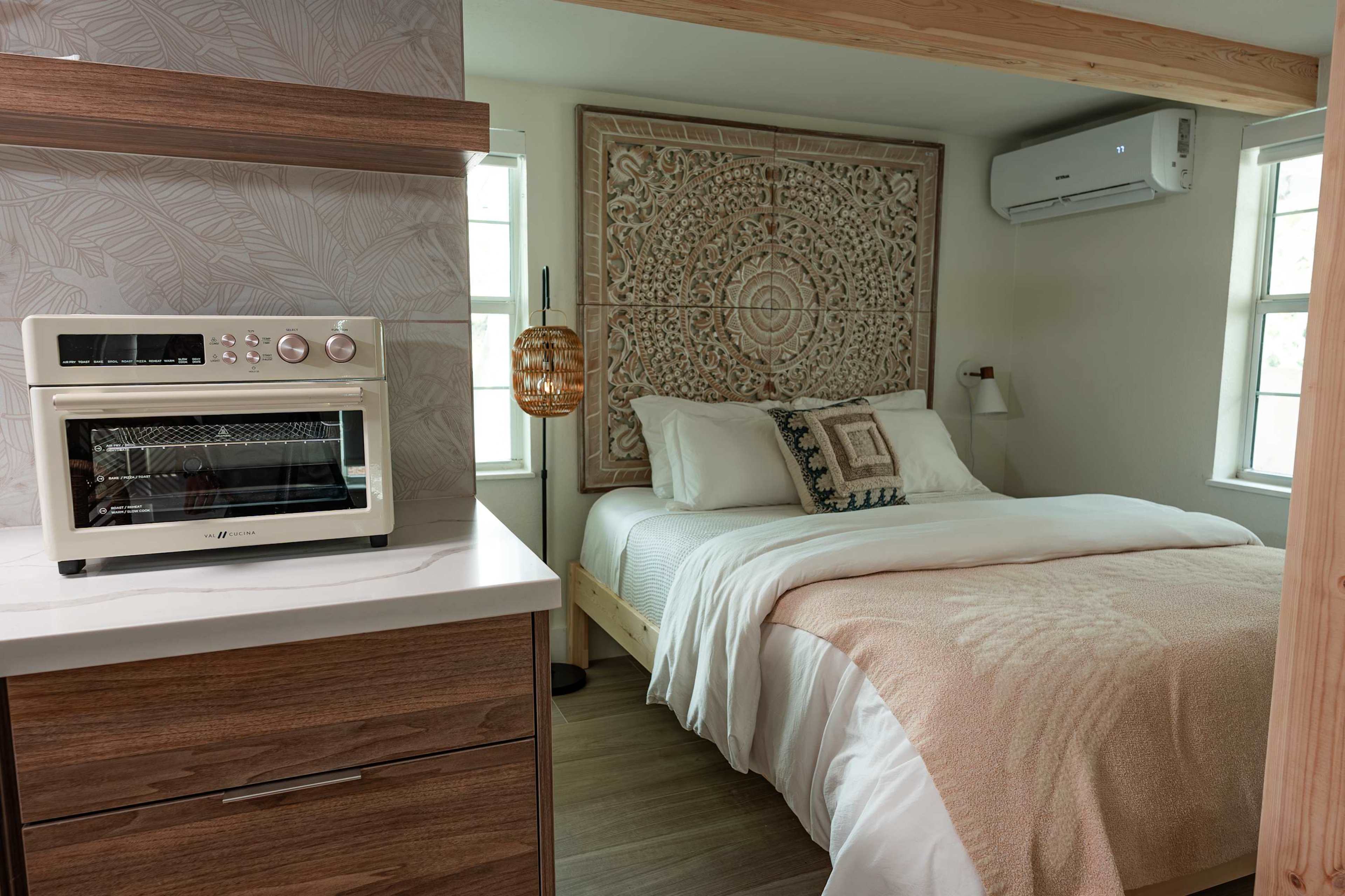 A cozy bedroom features a bed with a light-colored blanket, intricately carved wall decor, and an air conditioning unit, alongside a compact kitchen area with a countertop oven.