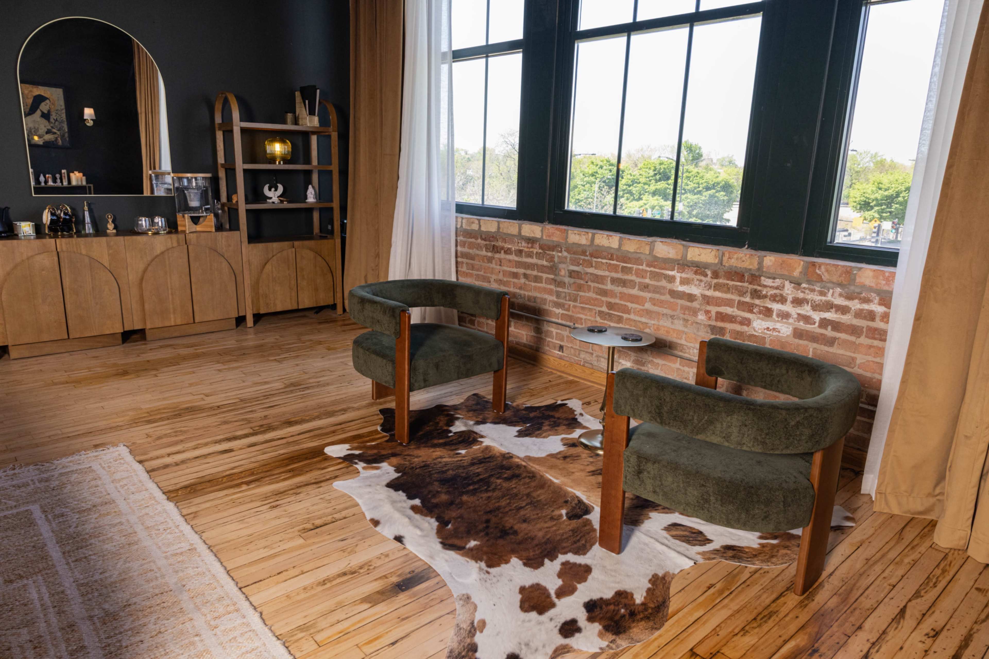 The image shows a spacious interior featuring two green armchairs positioned on a cowhide rug, with a wooden side table and large windows allowing natural light to enter the room.