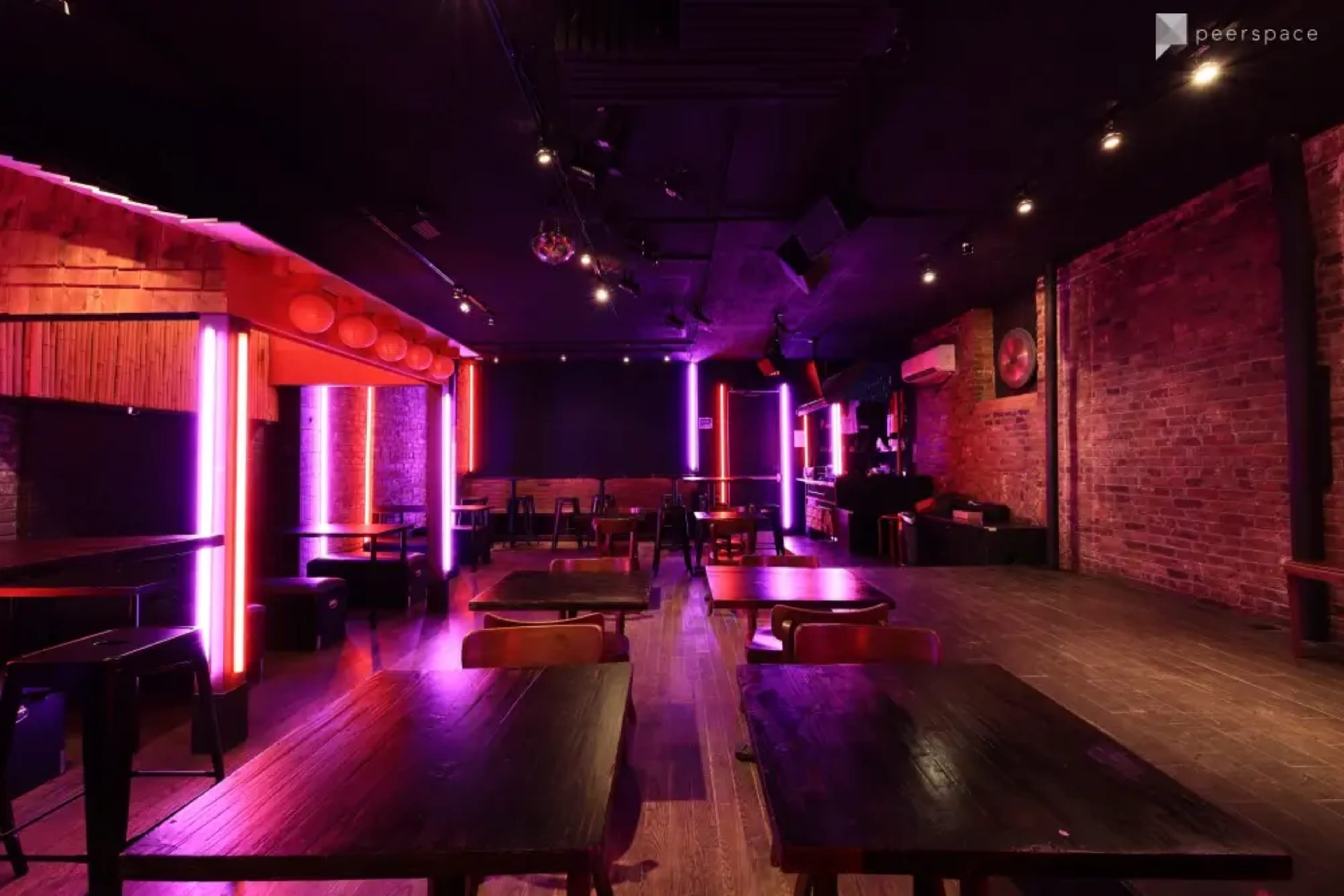The image shows a dimly lit interior of a lounge or nightclub featuring wooden tables and vibrant neon lighting along the walls.