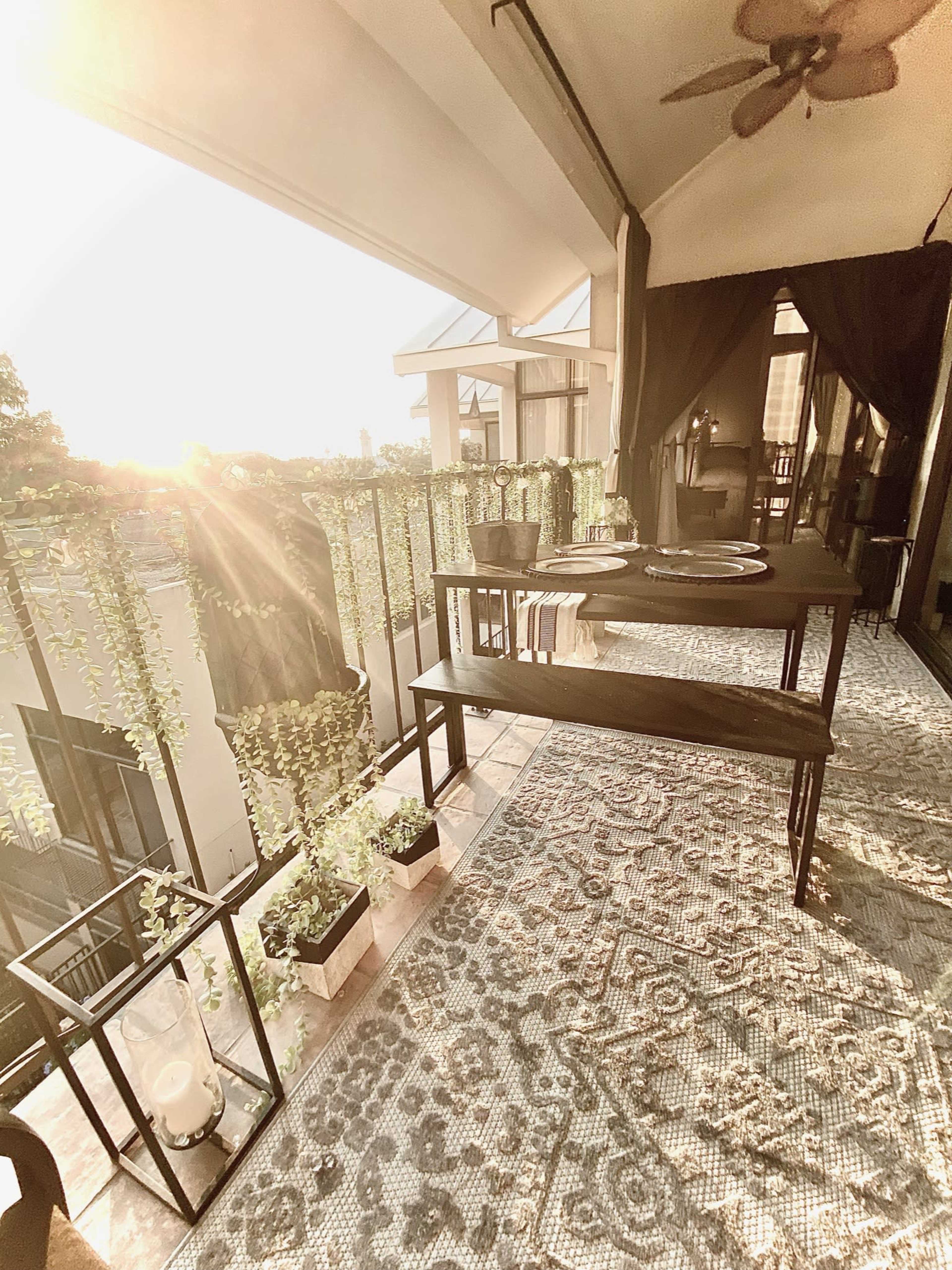 A balcony features a table set for dining, surrounded by potted plants and illuminated by the setting sun.