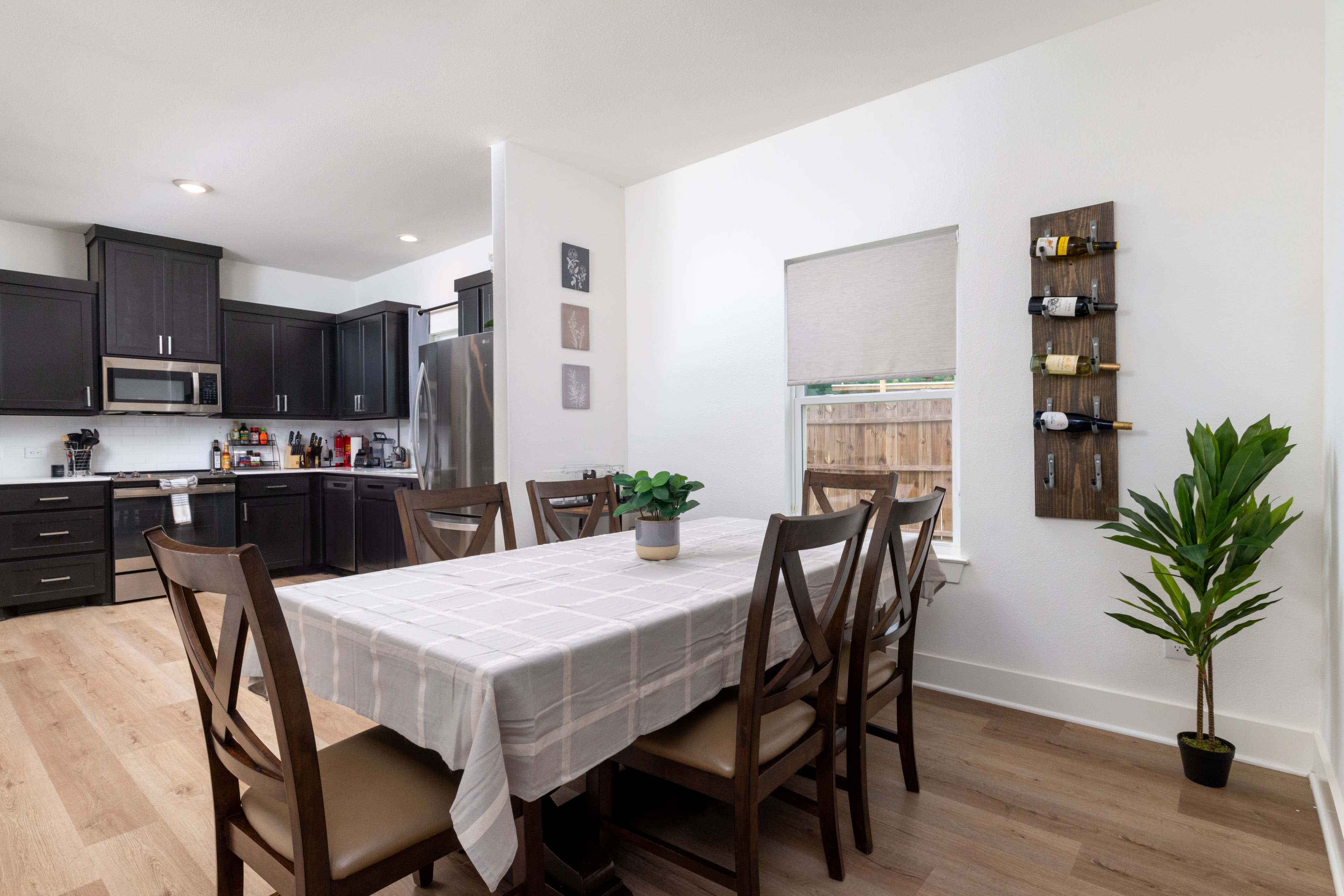 A modern kitchen and dining area features a large wooden table with a tablecloth, surrounded by chairs, while kitchen cabinets and appliances are visible in the background.