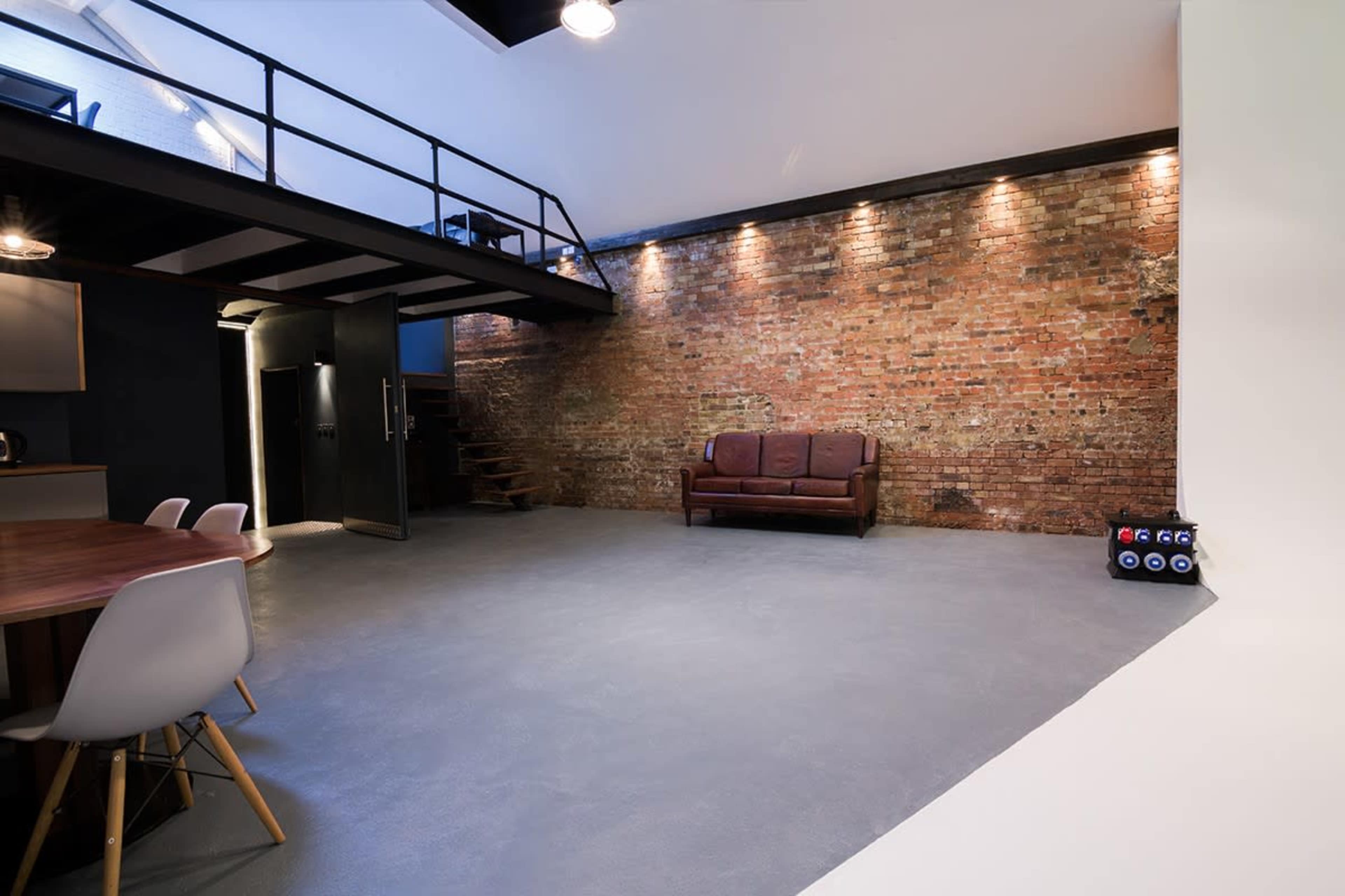 The image shows an industrial-style room with exposed brick walls, a leather couch, and a staircase leading to a loft area.