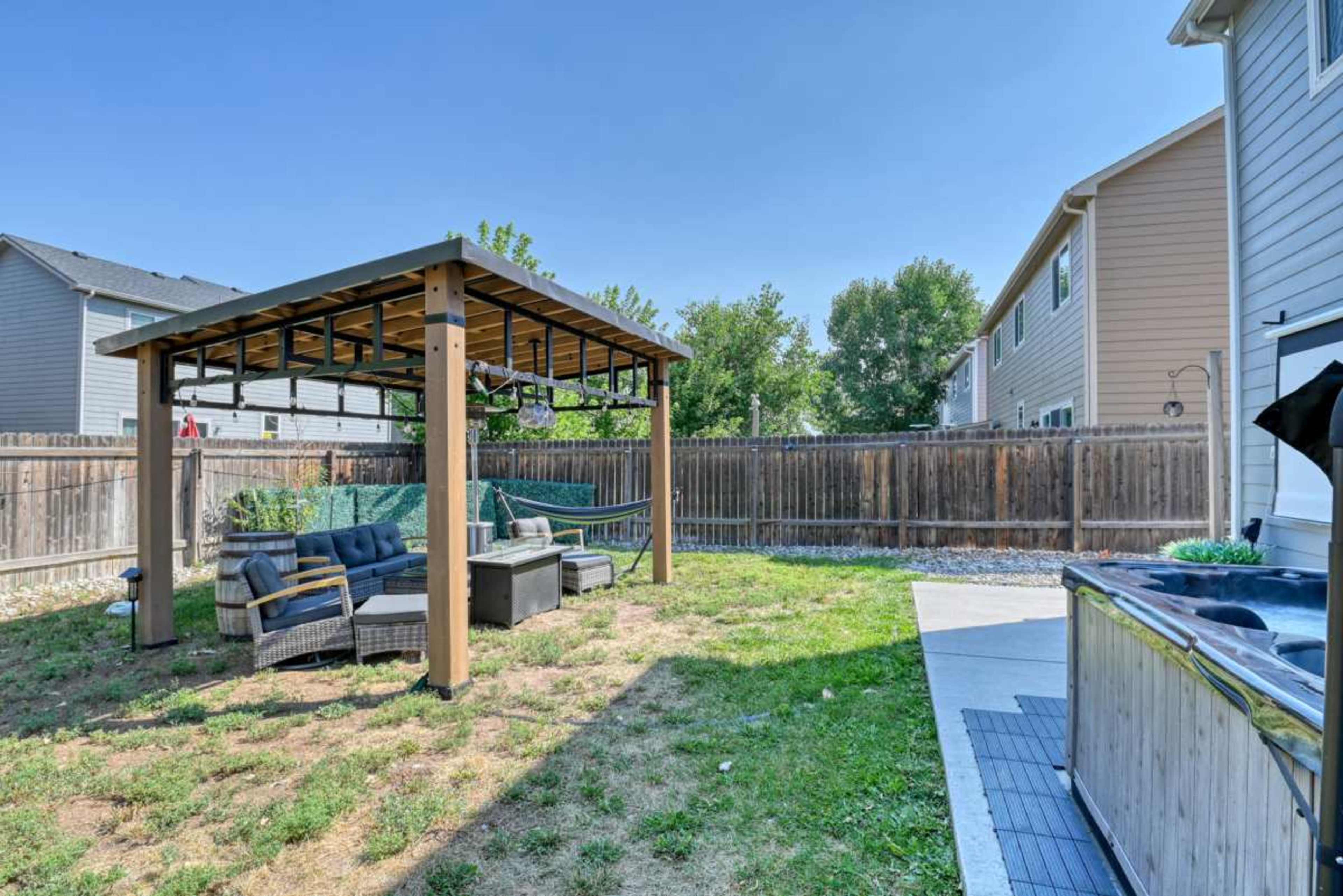 The image shows a backyard with a covered pergola containing seating, a grassy area, and a hot tub next to the house.