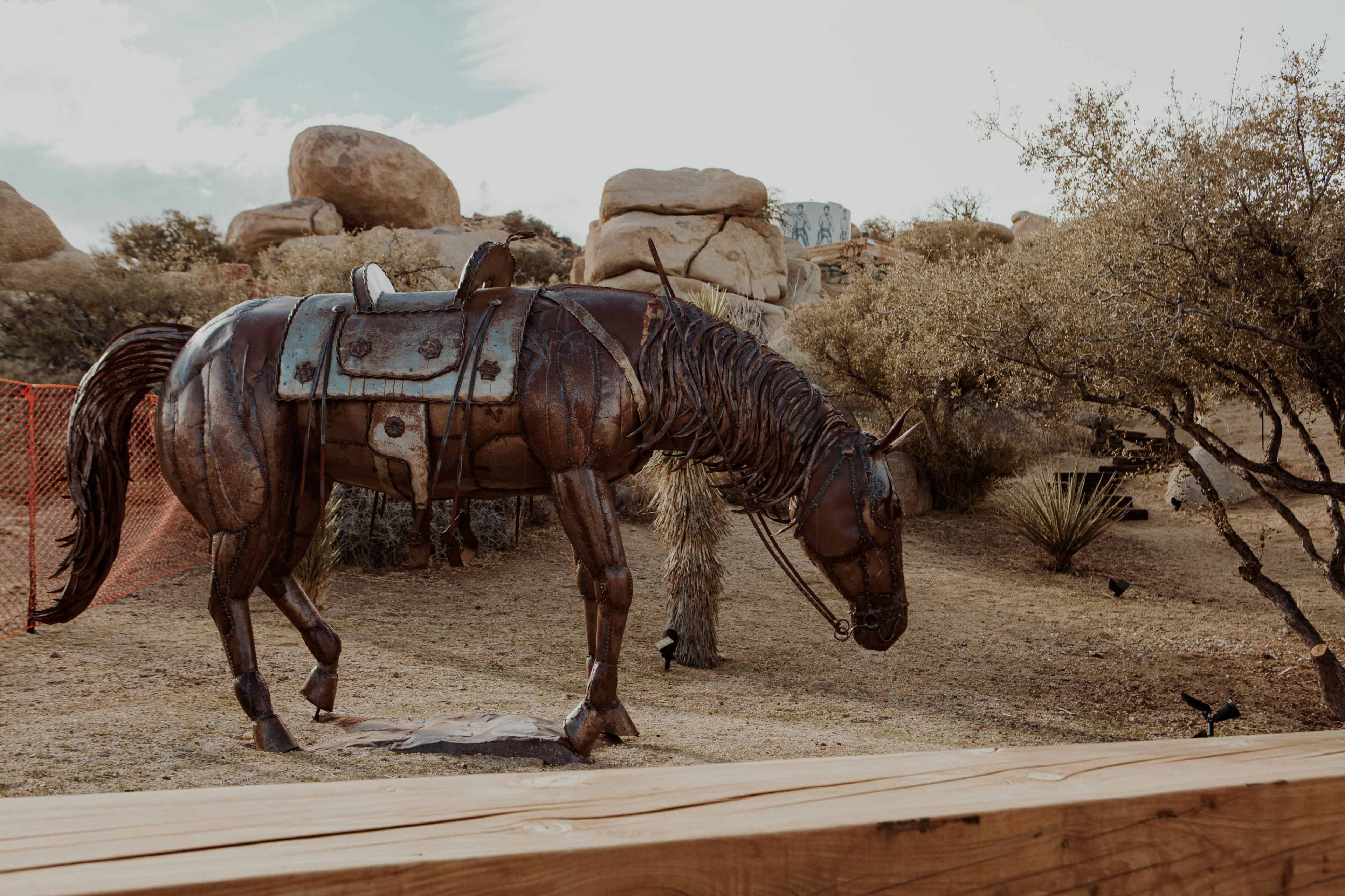 A metal sculpture of a horse stands in a desert landscape surrounded by rocks and sparse vegetation.