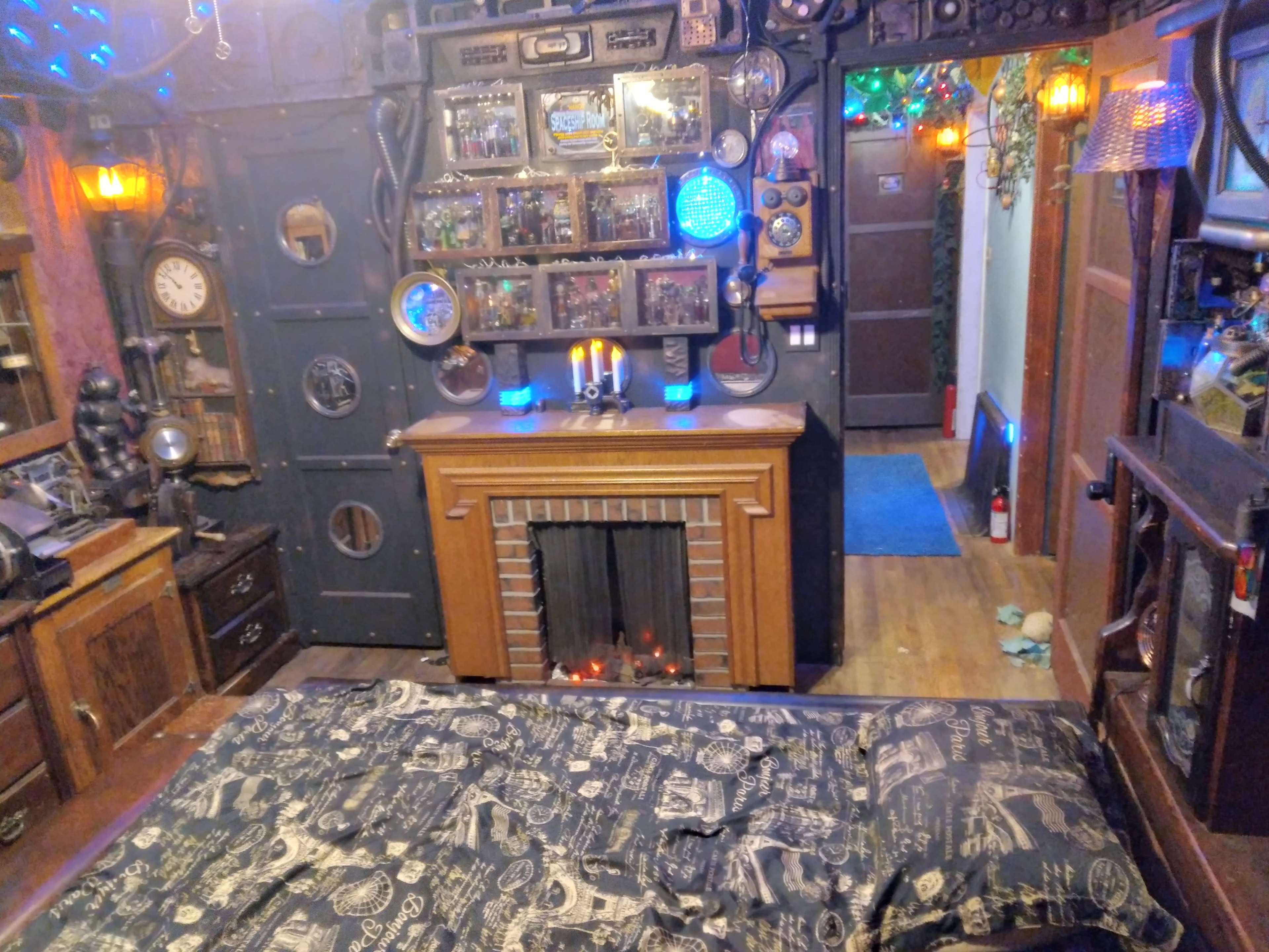 The room features a bed in front of a fireplace, surrounded by a wall adorned with various clocks, mirrors, and decorative items, illuminated by warm lighting.