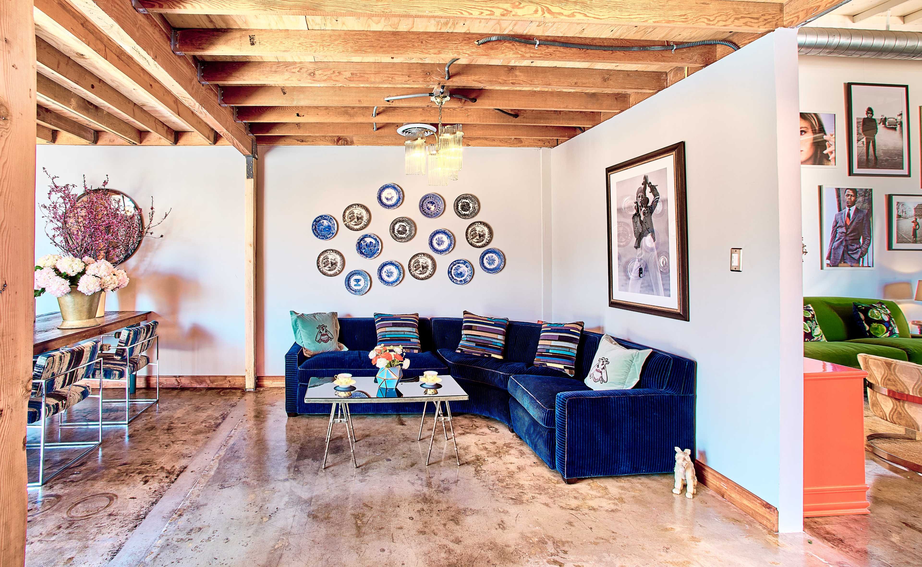 The scene features a modern living area with a blue sectional couch adorned with striped pillows, a round dining table, and decorative wall plates arranged in a circular pattern.