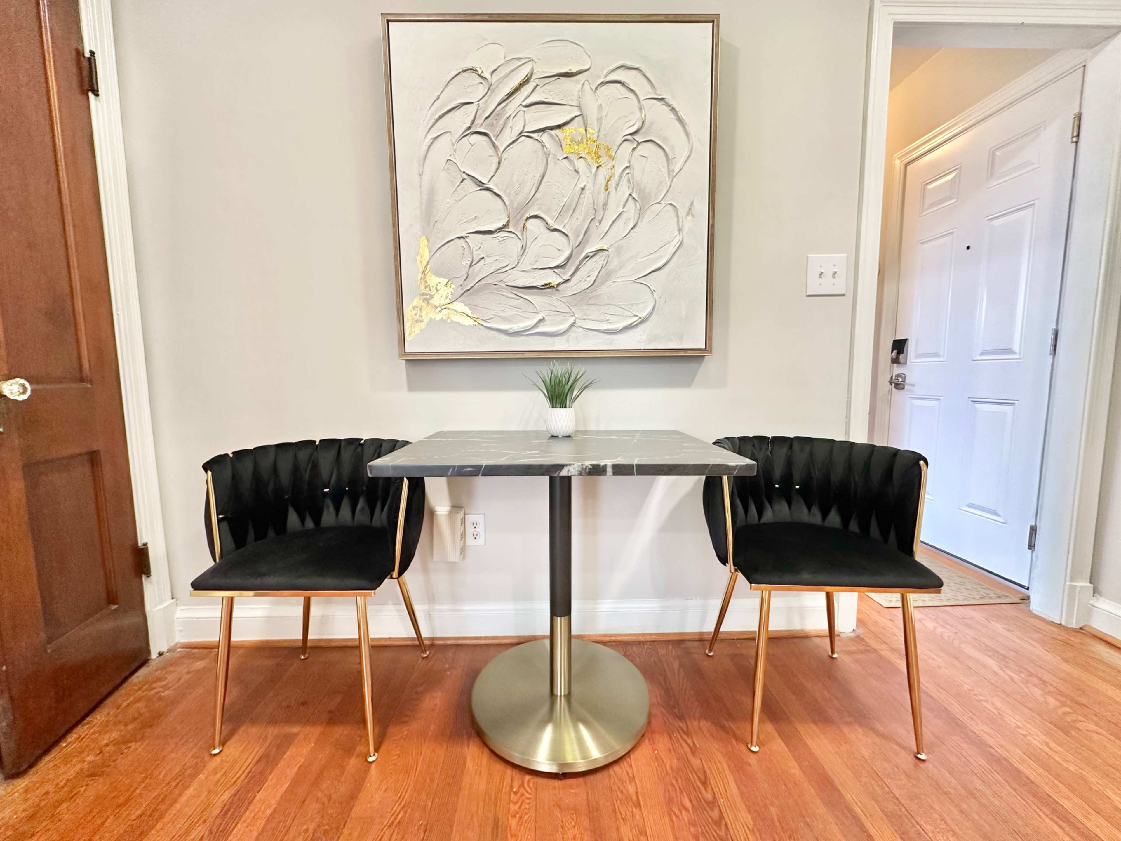 A small dining area features a round marble-top table with a metallic base and two black, ribbed chairs, set against a wall with a floral artwork.