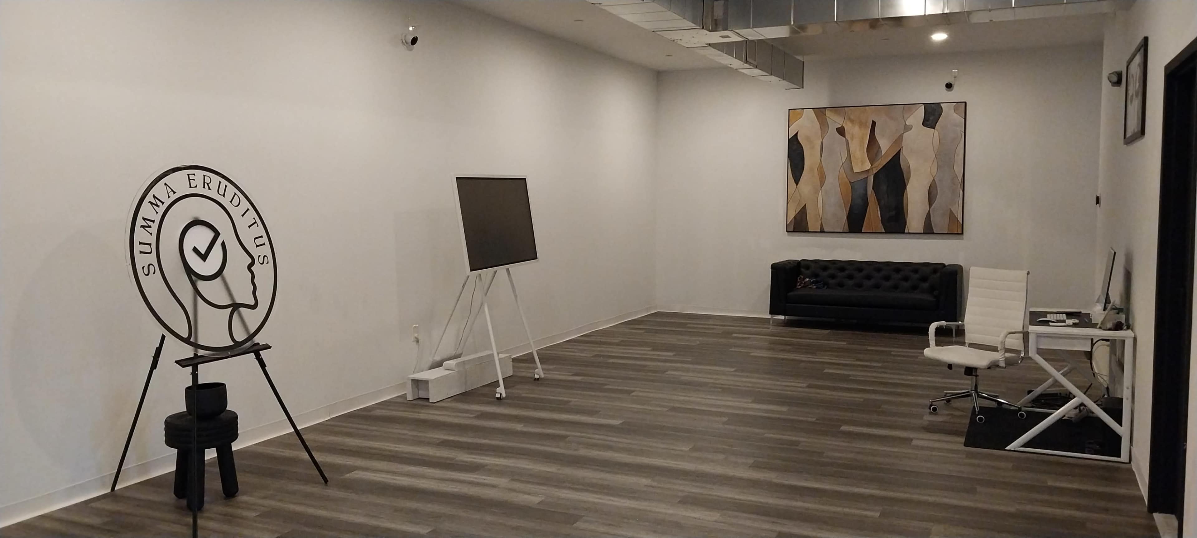 The image shows a spacious, minimalist room with wooden flooring, featuring a black couch, an easel with a blank board, and an abstract artwork on the wall.
