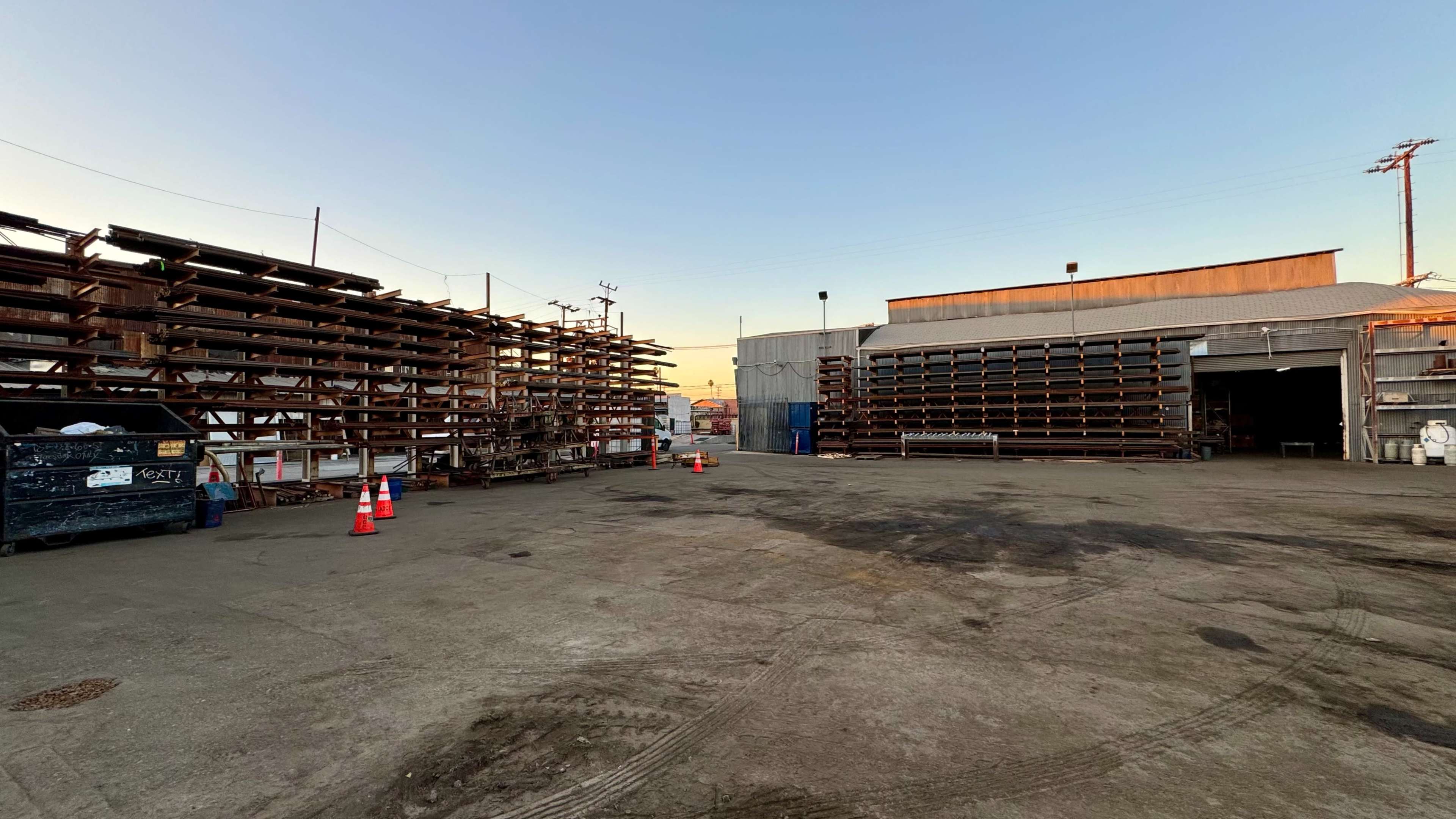 The image shows a storage area with stacked metal pipes on the left and a warehouse building on the right, under a clear sky at dusk.