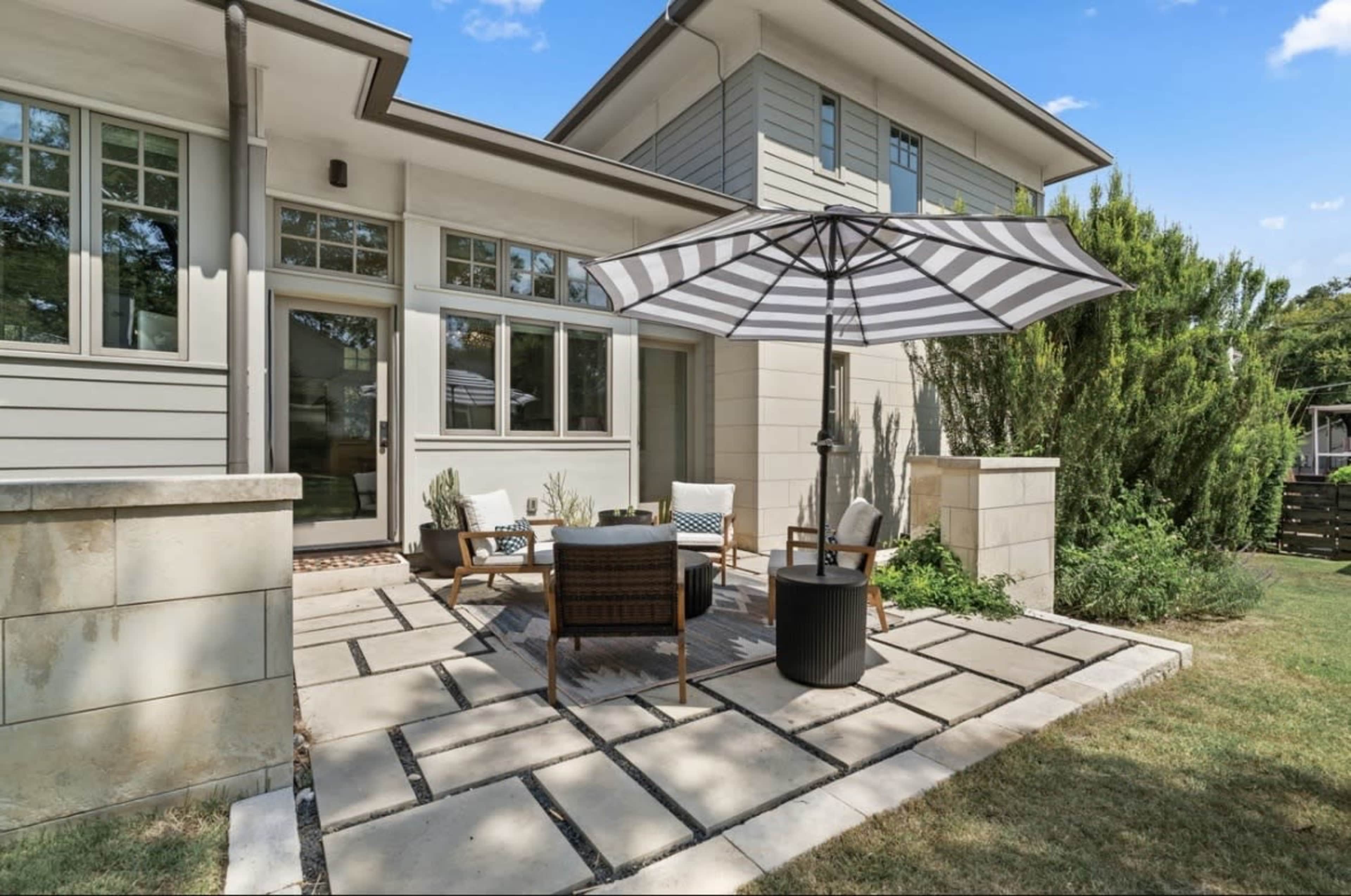 A small patio area features a table and chairs under a striped umbrella, surrounded by landscaped grass and plants.