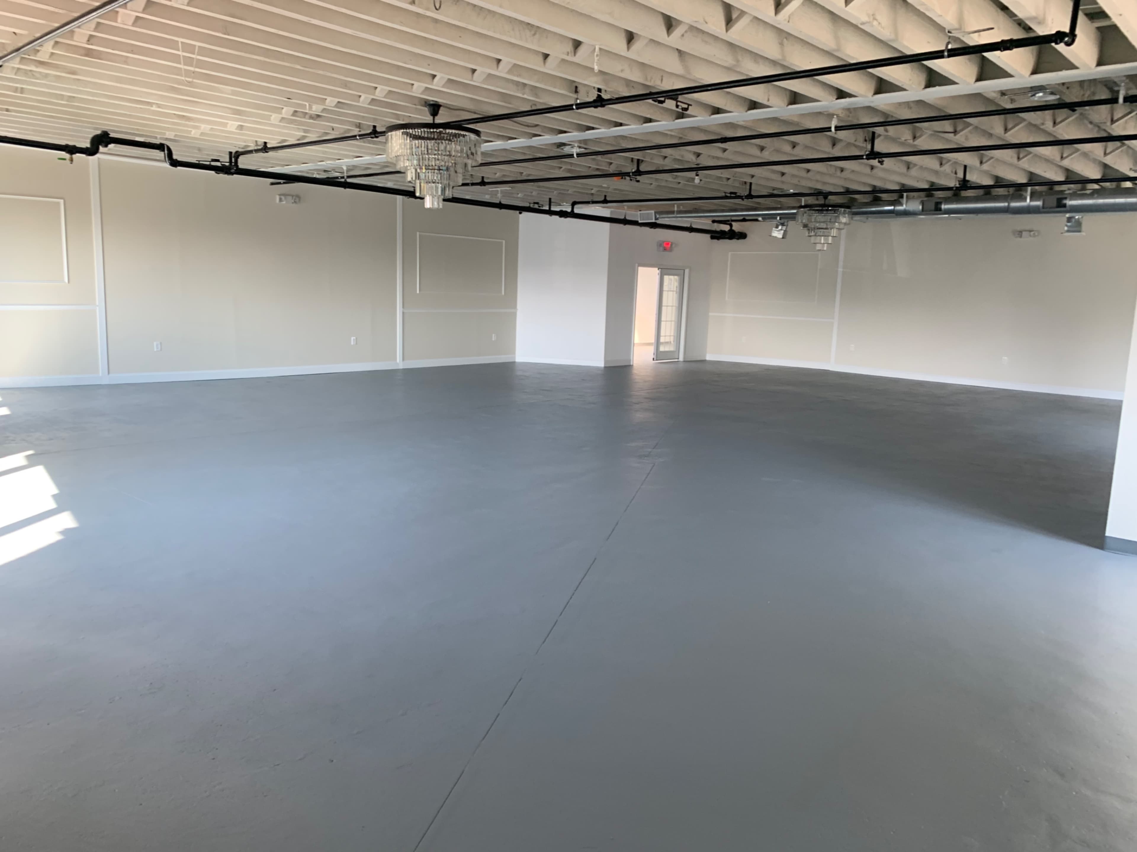 A large, empty room with a gray concrete floor and exposed ceiling beams, featuring several light fixtures and a door at one end.