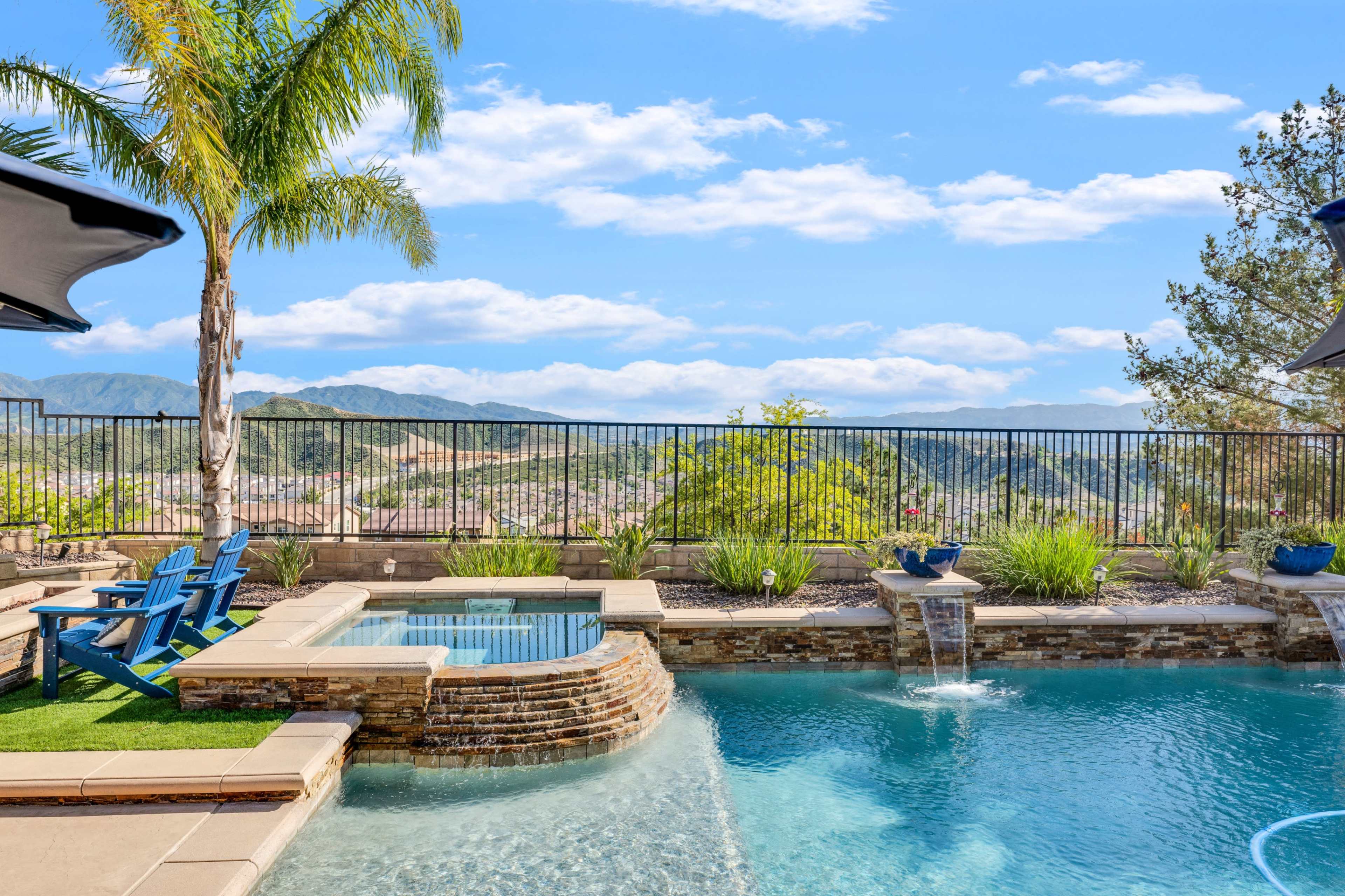 The image shows a landscaped pool area with a spa, blue lounge chairs, and a panoramic view of mountains and sky.