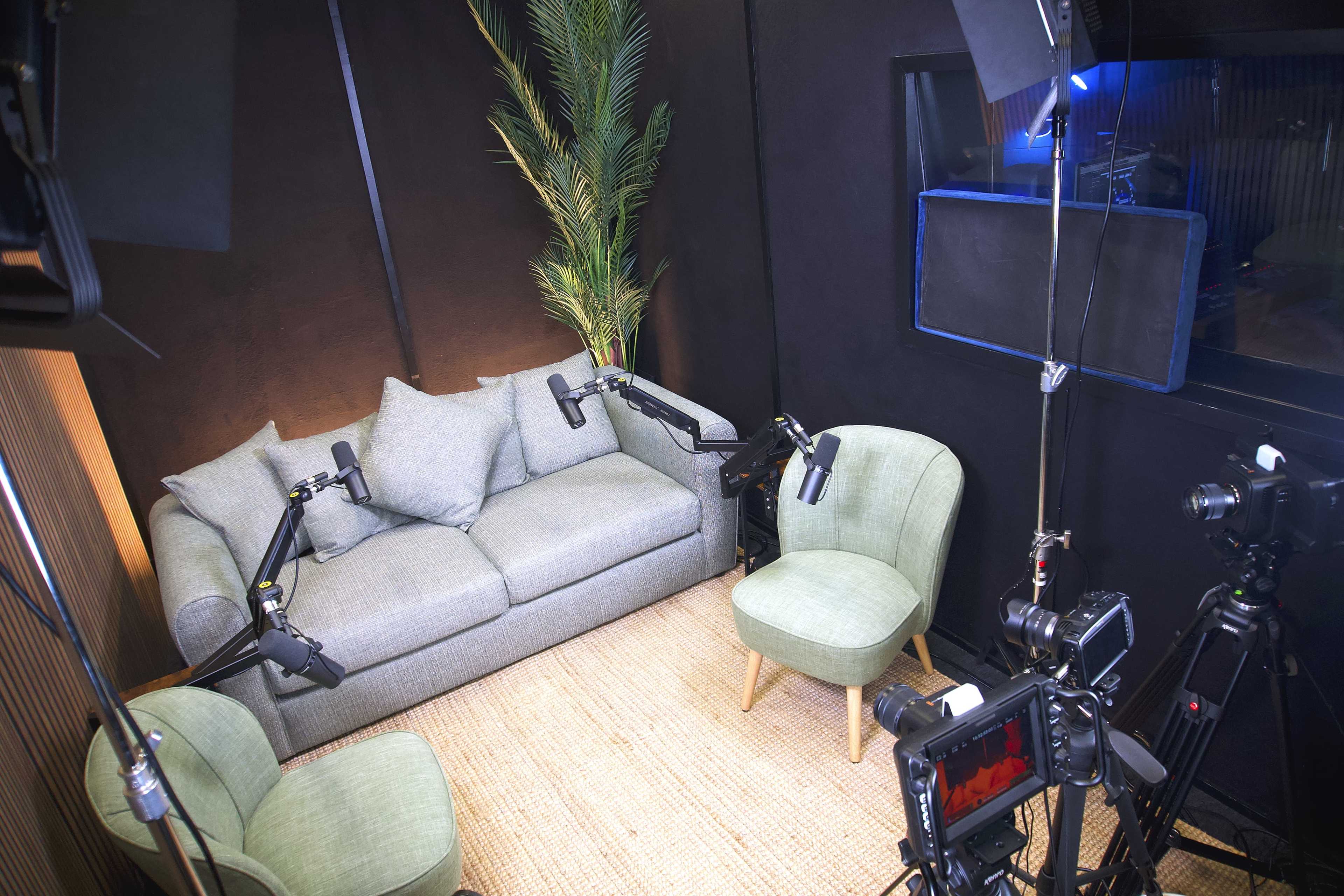 The image shows a small, modern recording studio setup with a gray couch, two green chairs, and multiple cameras and lights positioned around the seating area.