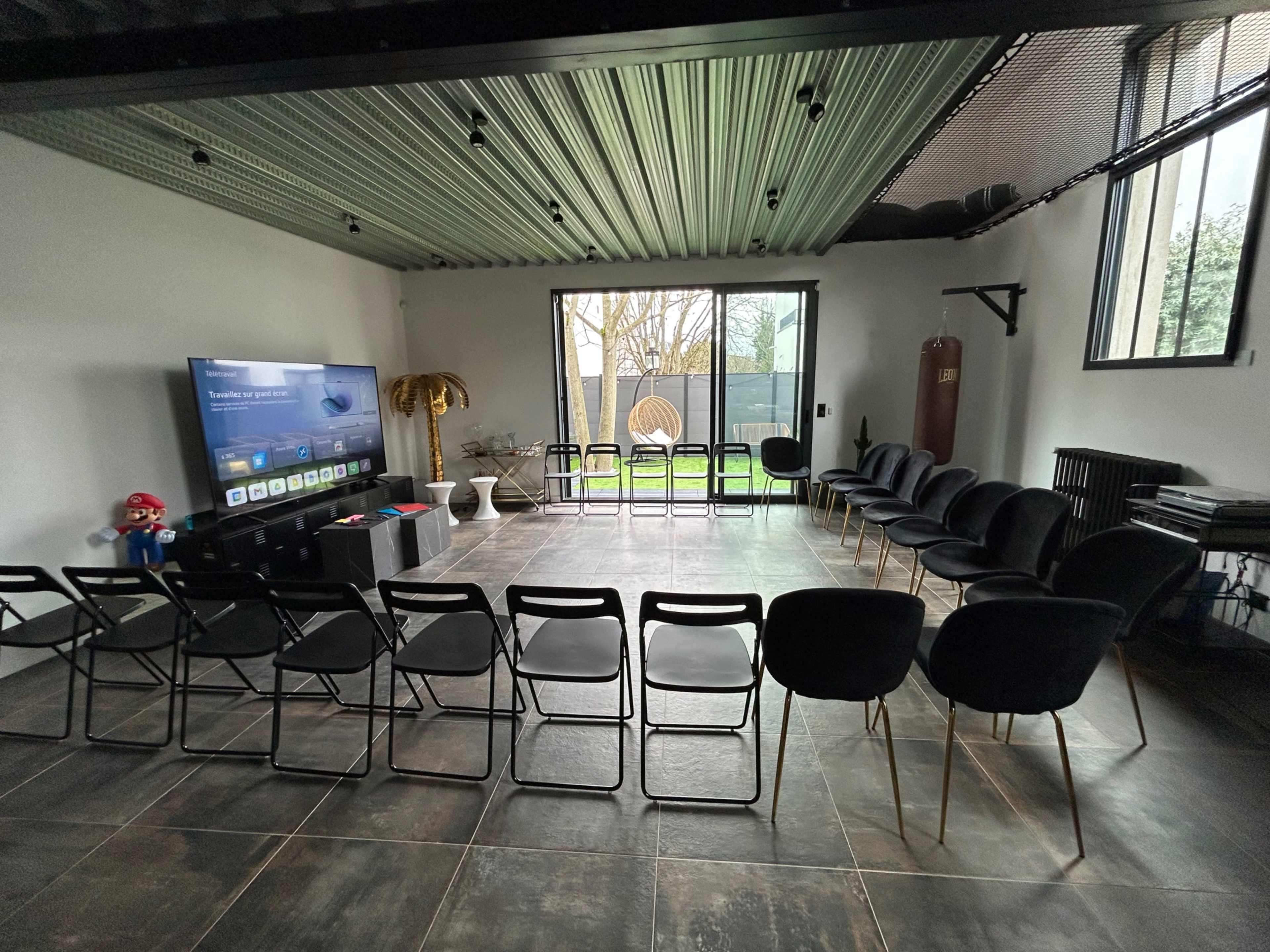 The image shows a meeting room with rows of black chairs facing a television and a large window overlooking a garden area.