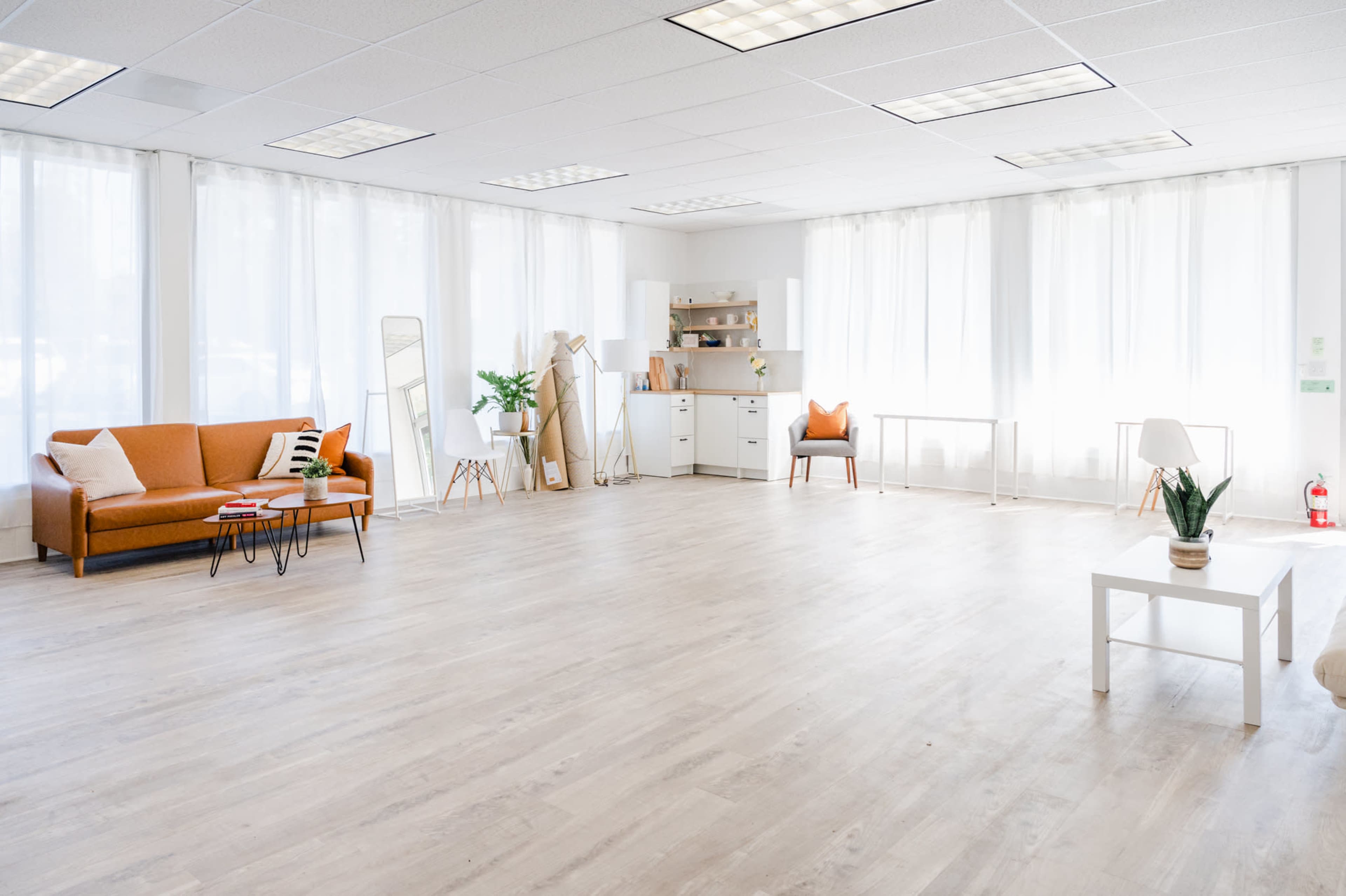 The image shows a spacious, bright room with large windows, featuring a brown sofa, a white table, and a minimalist decor arrangement.