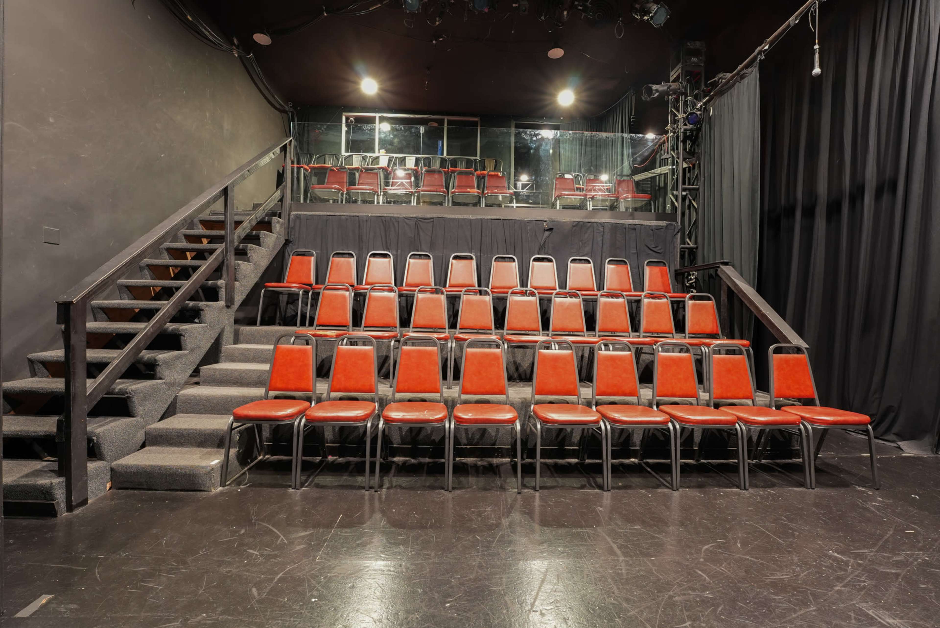 The image shows a small theater with a set of red chairs arranged in rows, situated in front of a stage and a staircase leading to a higher seating area.