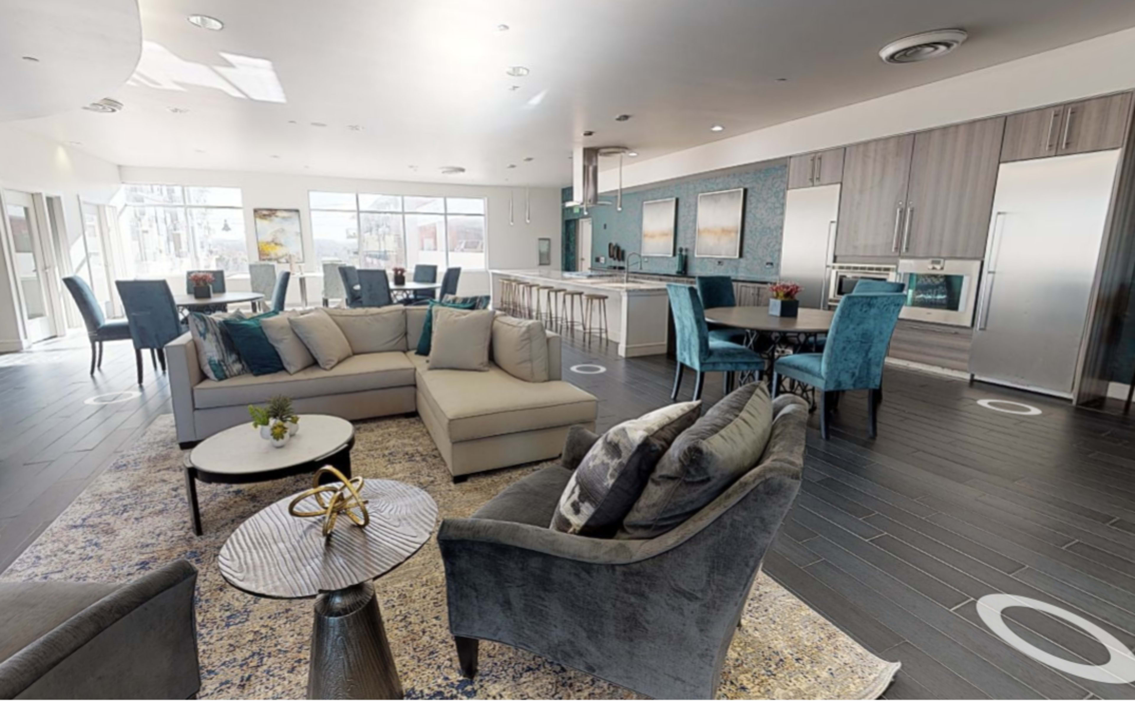 A modern communal lounge area with comfortable seating, a kitchen space, and dining tables arranged around large windows.