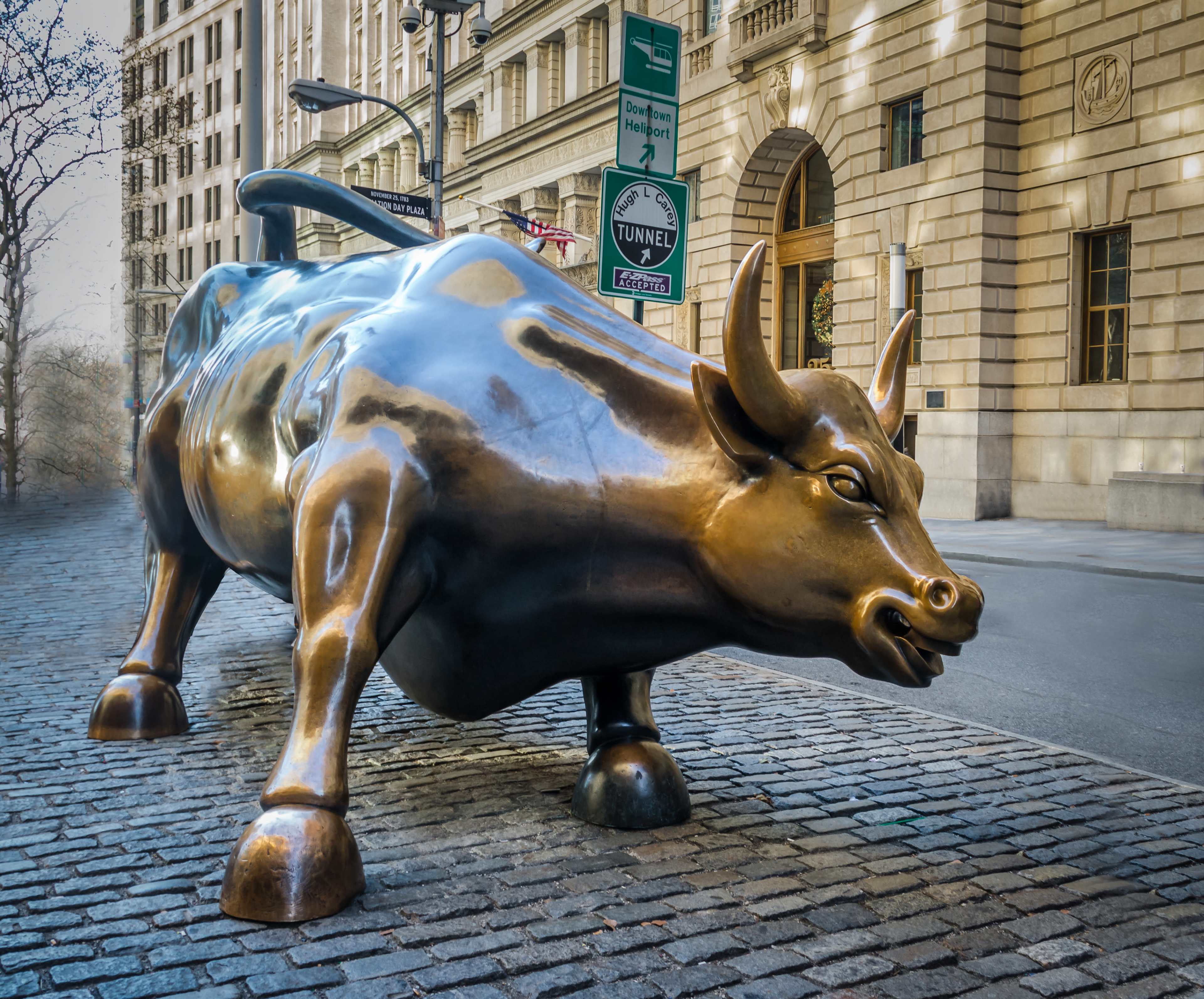 A large bronze bull sculpture positioned on a cobblestone street with tall buildings in the background.