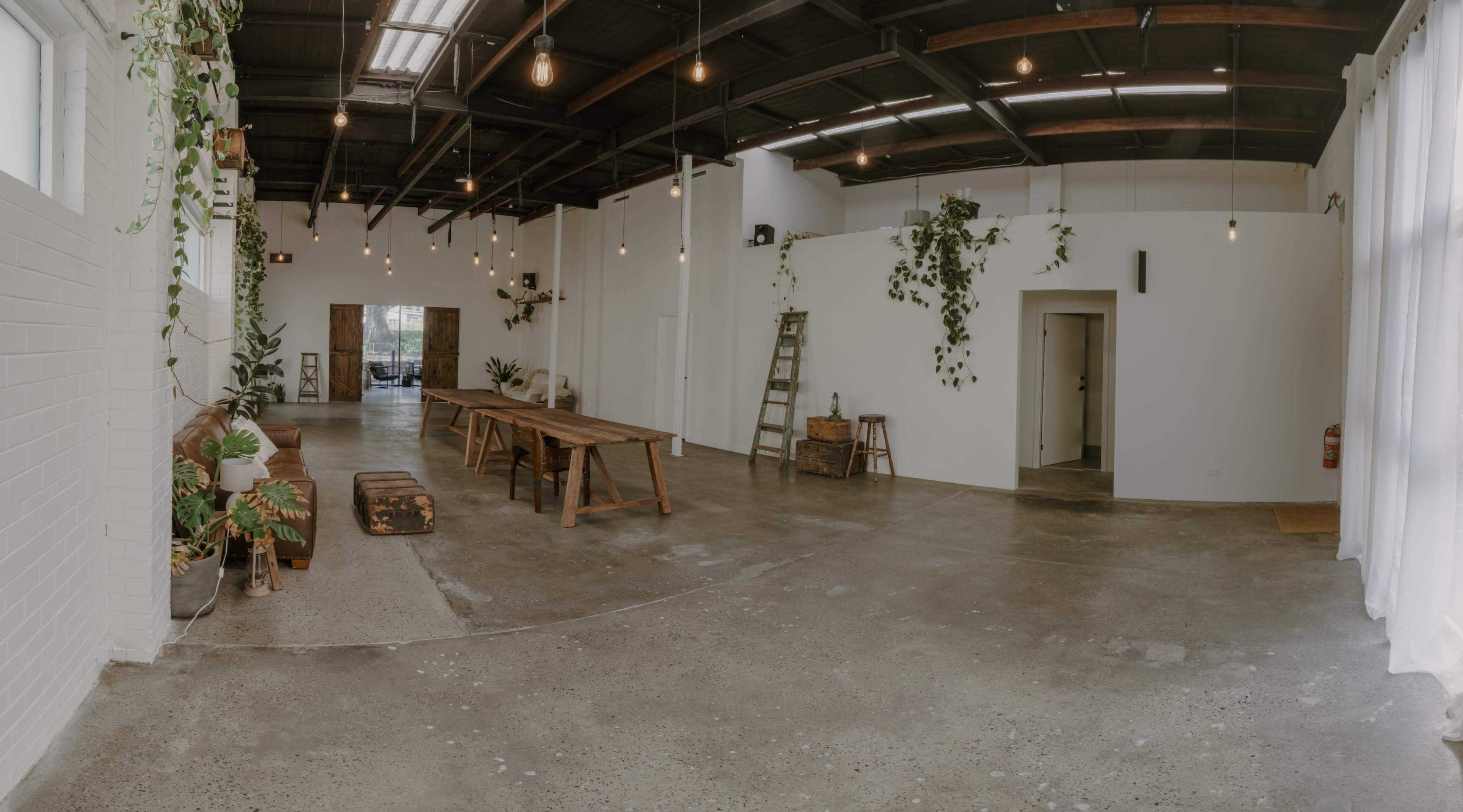 An industrial-style room features a long wooden table, hanging lights, and greenery, with concrete floors and white walls.